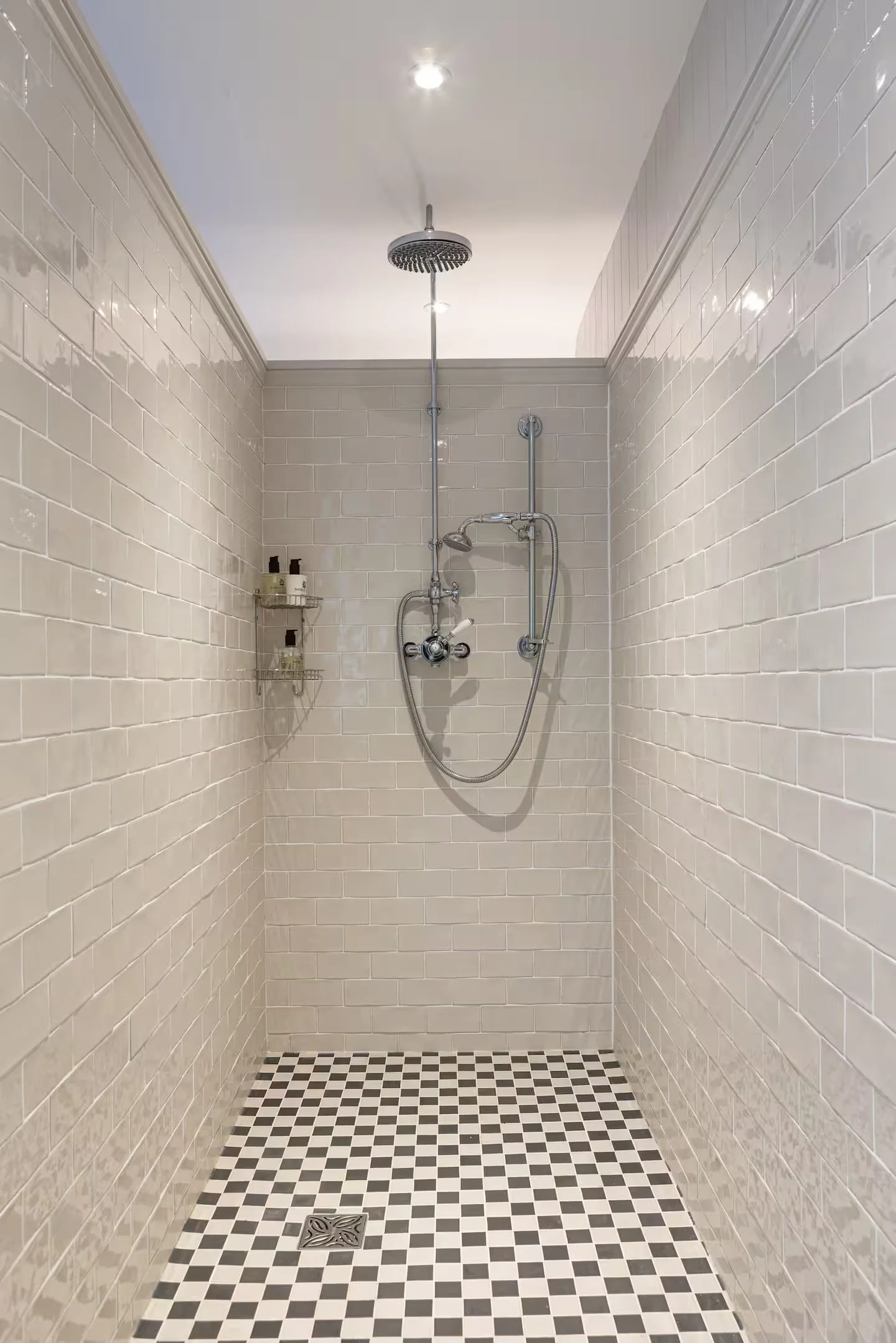 Showers with beige subway tiles on the walls, a black-and-white checkered floor, a ceiling drain, and a showerhead with a handheld hose, along with some toiletries on a metal shelf.
