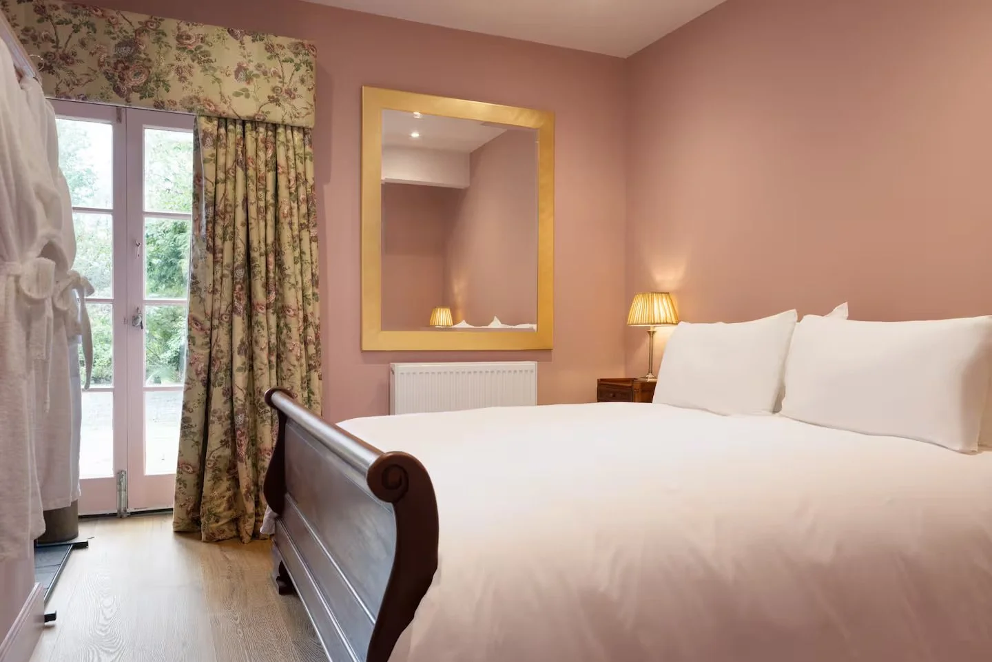 A cozy bedroom with pink walls, a large bed with white linens and pillows, floral curtains, a mirror with a gold frame, a wooden side table with a lamp, and a glass door leading outside.