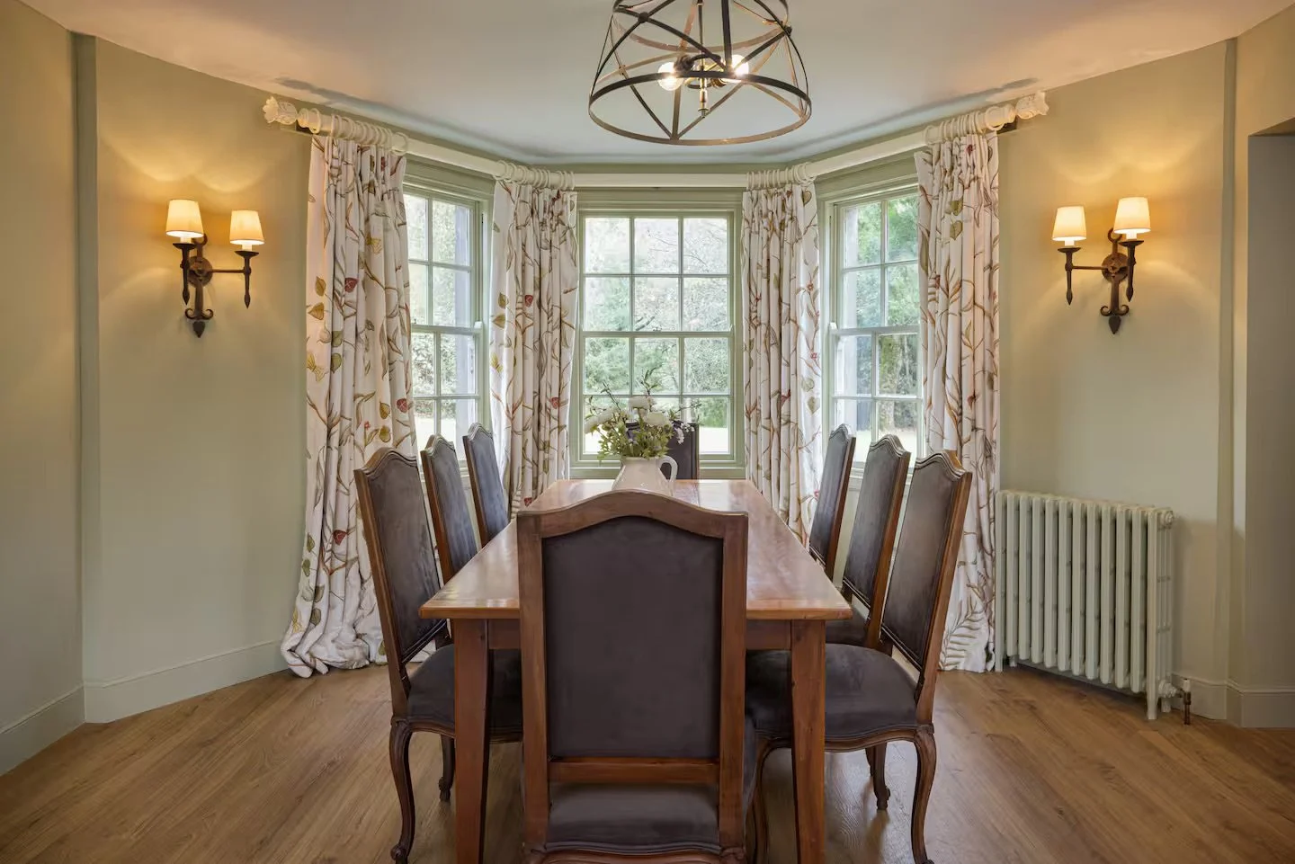 Dining room with a wooden table, six matching chairs, floral curtains, a centerpiece with flowers, window with greenery outside, wall sconces with lights, and a chandelier