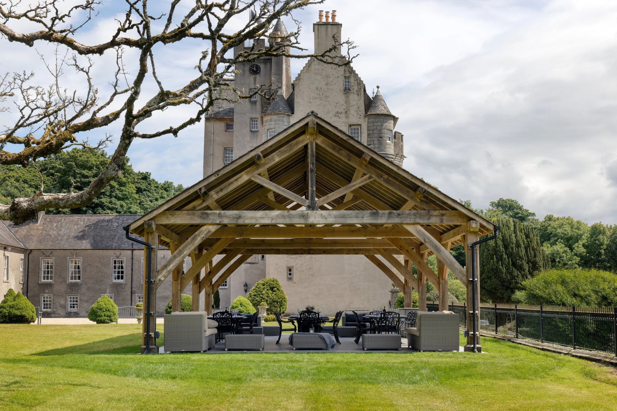 An outdoor patio setup with chairs and sofas under a wooden pergola in front of a large historic castle with stone walls, towers, and a clock on one of its towers. A leafless tree is in the foreground and the sky is partly cloudy.