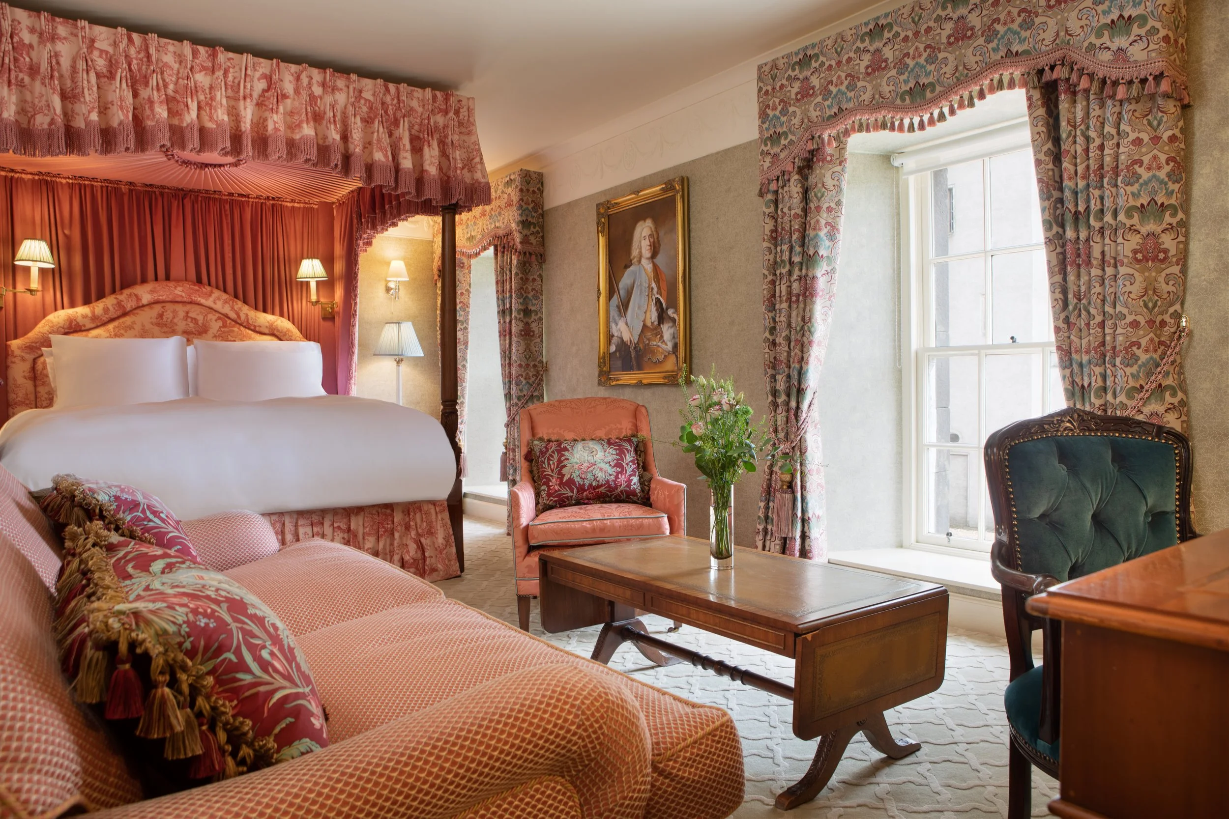 A luxurious, vintage-style bedroom with pink and floral decor, plush furniture, lace curtains, and a portrait of a historical figure on the wall.