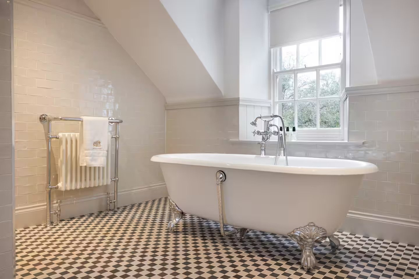 A vintage clawfoot bathtub in a bright bathroom with large window, white tiled walls, checkered black and white floor, and radiator with towel.