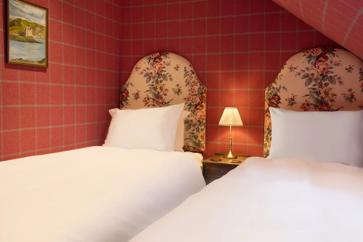 Two beds with floral-patterned headboards in a room with pink plaid wallpaper, a small wooden nightstand with a lamp, and a framed landscape painting on the wall.