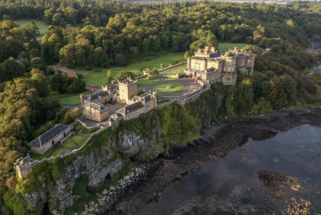Aerial view of a historic castle on a rocky coastline surrounded by lush green trees and grass, overlooking the water.