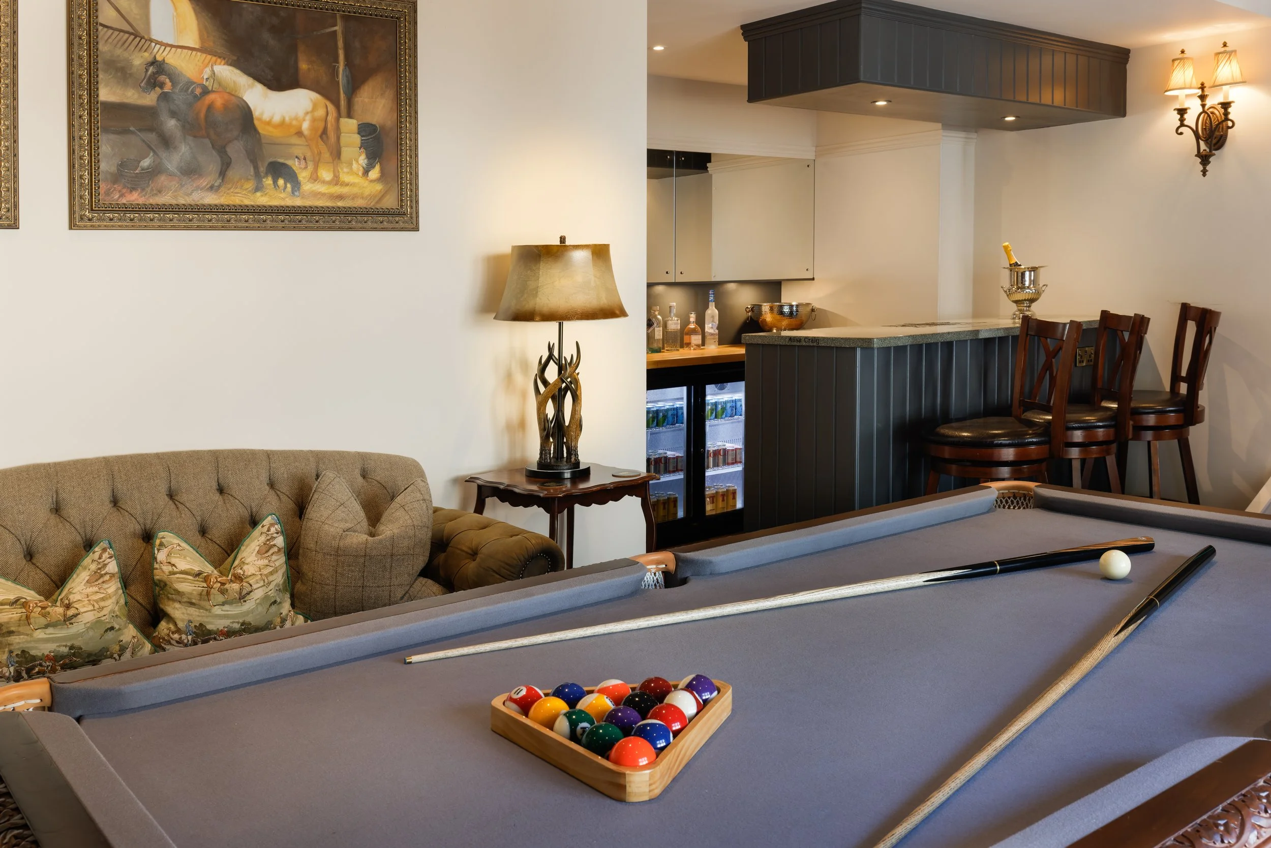 Indoor entertainment area with pool table set up for billiards, cue sticks, a wooden tray of pool balls, a bar counter with bottles, a vintage silver punch bowl, a bar stool, modern lighting, a framed painting of horses on the wall, a lamp with a decorative base, and a sofa with floral throw pillows.
