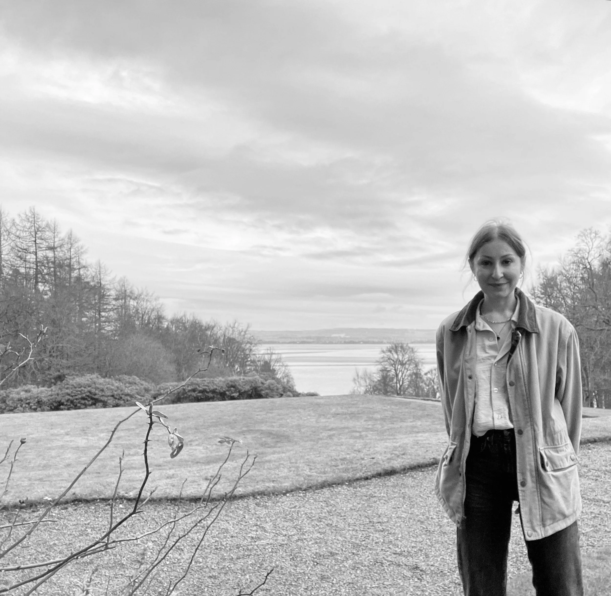 A woman with light-colored hair standing outdoors near a small branch with leaves, with a lake, leafless trees, and cloudy sky in the background.