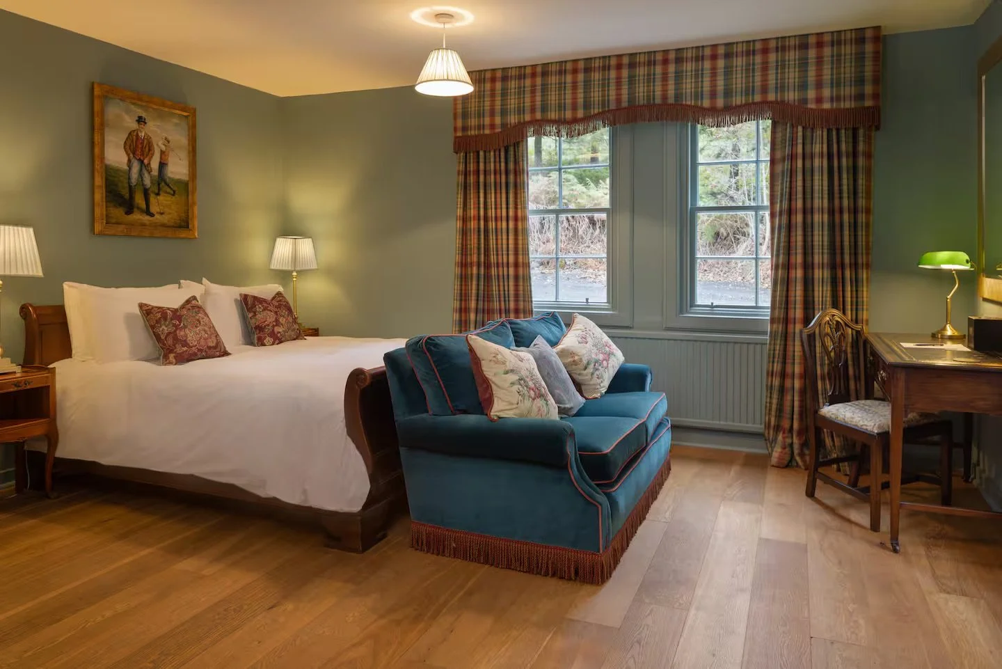 A cozy bedroom with a large bed, wooden nightstand, and two lamps on either side. There is a vintage-style blue velvet sofa with floral pillows in front of windows with plaid curtains. A picture of two men playing golf hangs on the green-painted wall. Wooden flooring completes the room's warm ambiance.
