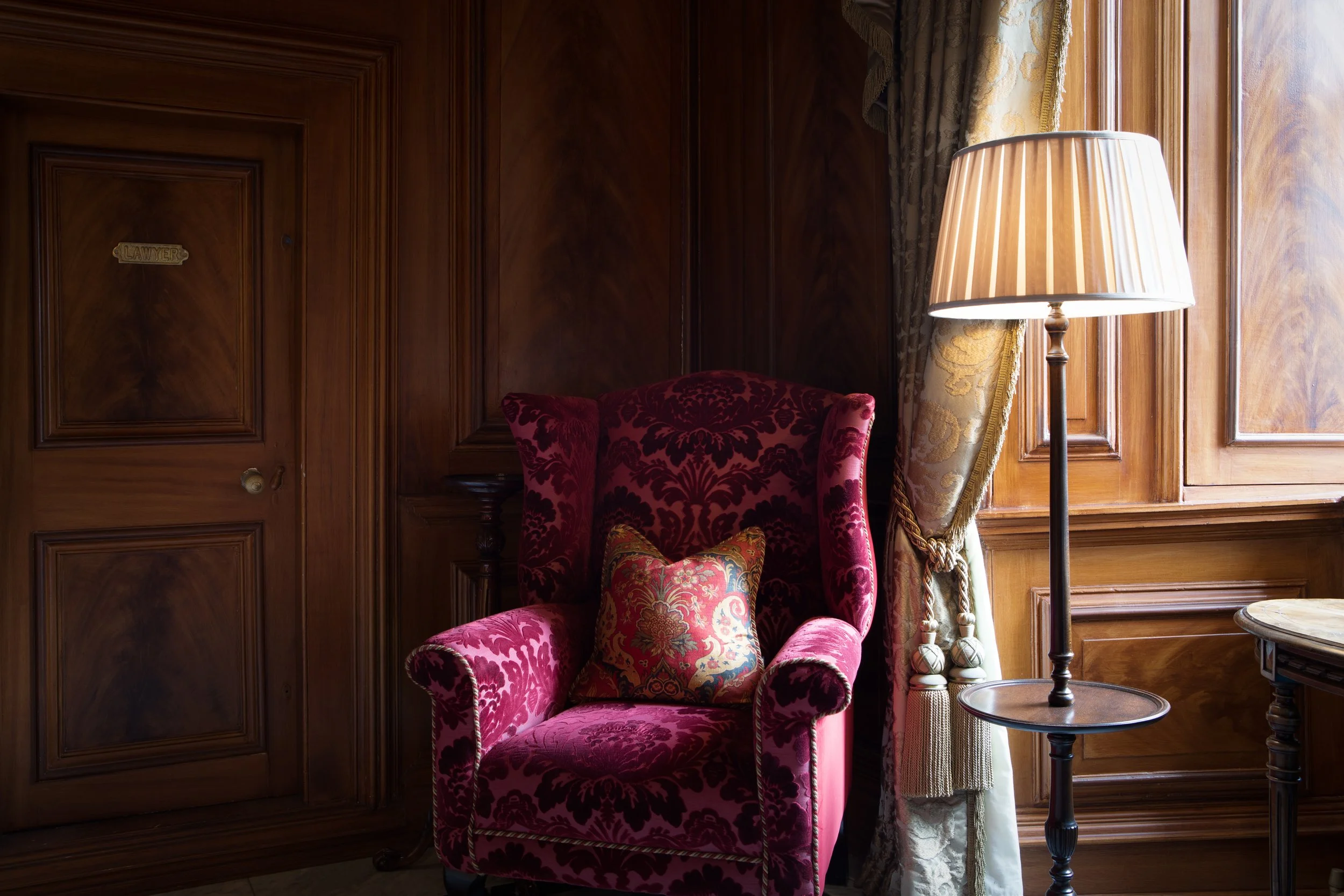 A luxurious red velvet armchair with floral pillows, illuminated by a nearby floor lamp, in a wood-paneled room with ornate curtains.