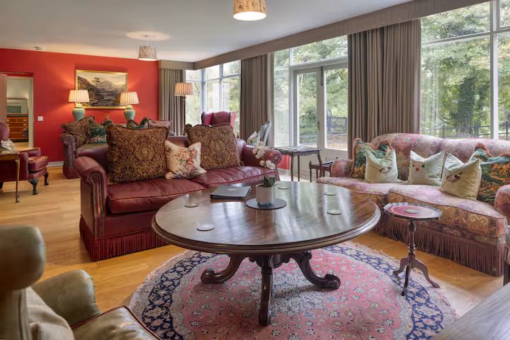 Living room with a large oval wooden coffee table, upholstered sofas with multiple decorative pillows, hardwood floors, and large windows with curtains showing greenery outside.