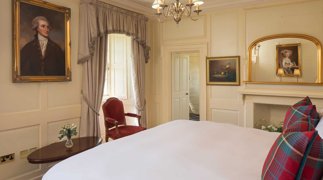 A bedroom with cream-colored walls, a window with drapes, a wooden chair, a side table with a flower vase, and framed portraits on the walls.