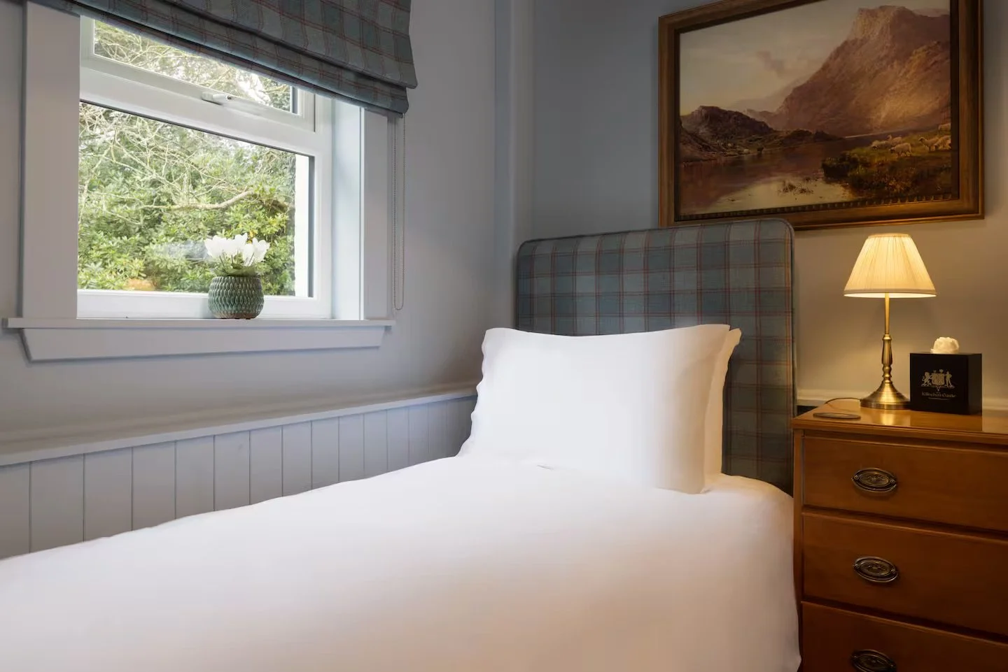 A cozy bedroom with a single bed, white bedding, and a plaid headboard, beside a wooden nightstand with a lamp, a framed photo, and a piece of rock. There is a window with a vase of flowers on the sill, and a landscape painting on the wall.