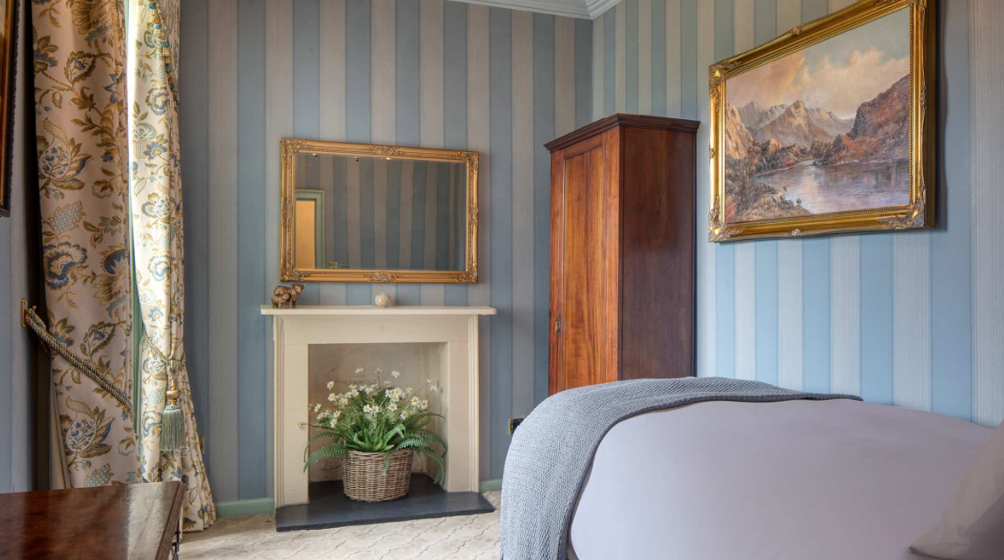 Interior of a bedroom with blue and gold curtains, framed paintings, a white fireplace with flowers, a wooden wardrobe, and a bed with gray bedding.