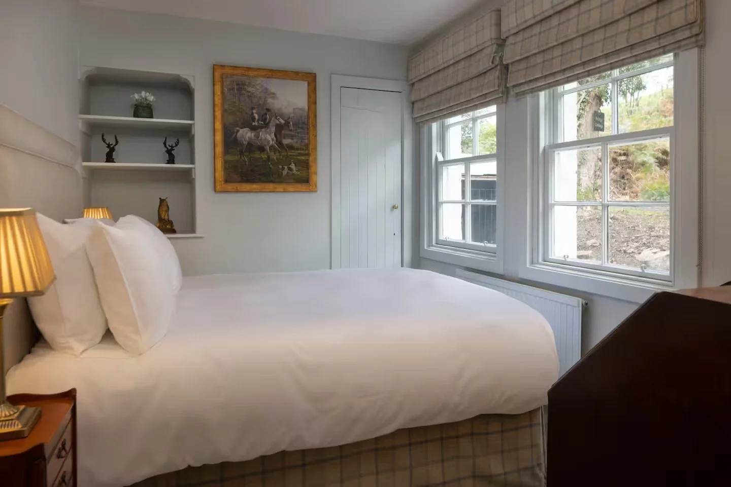 A cozy bedroom with a large bed, white bedding, and pillows. Beside the bed, there are two gold-striped lamps on wooden nightstands. The wall behind the bed features built-in shelves with decorative items, a framed painting of a person riding a horse, and a small carved animal figure. Large windows with beige blinds let in natural light and provide a view of trees outside.