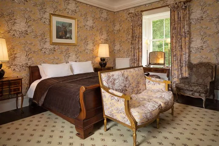 A bedroom with a wooden bed, floral wallpaper, a sofa with floral upholstery, a window with floral curtains, and a writing desk with a chair.
