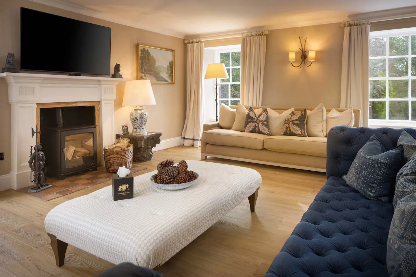 A cozy living room with beige walls, large windows with white curtains, a beige sofa with decorative pillows, a navy blue tufted armchair with patterned pillows, a white upholstered ottoman with pinecones, a fireplace with a large flat-screen TV moun