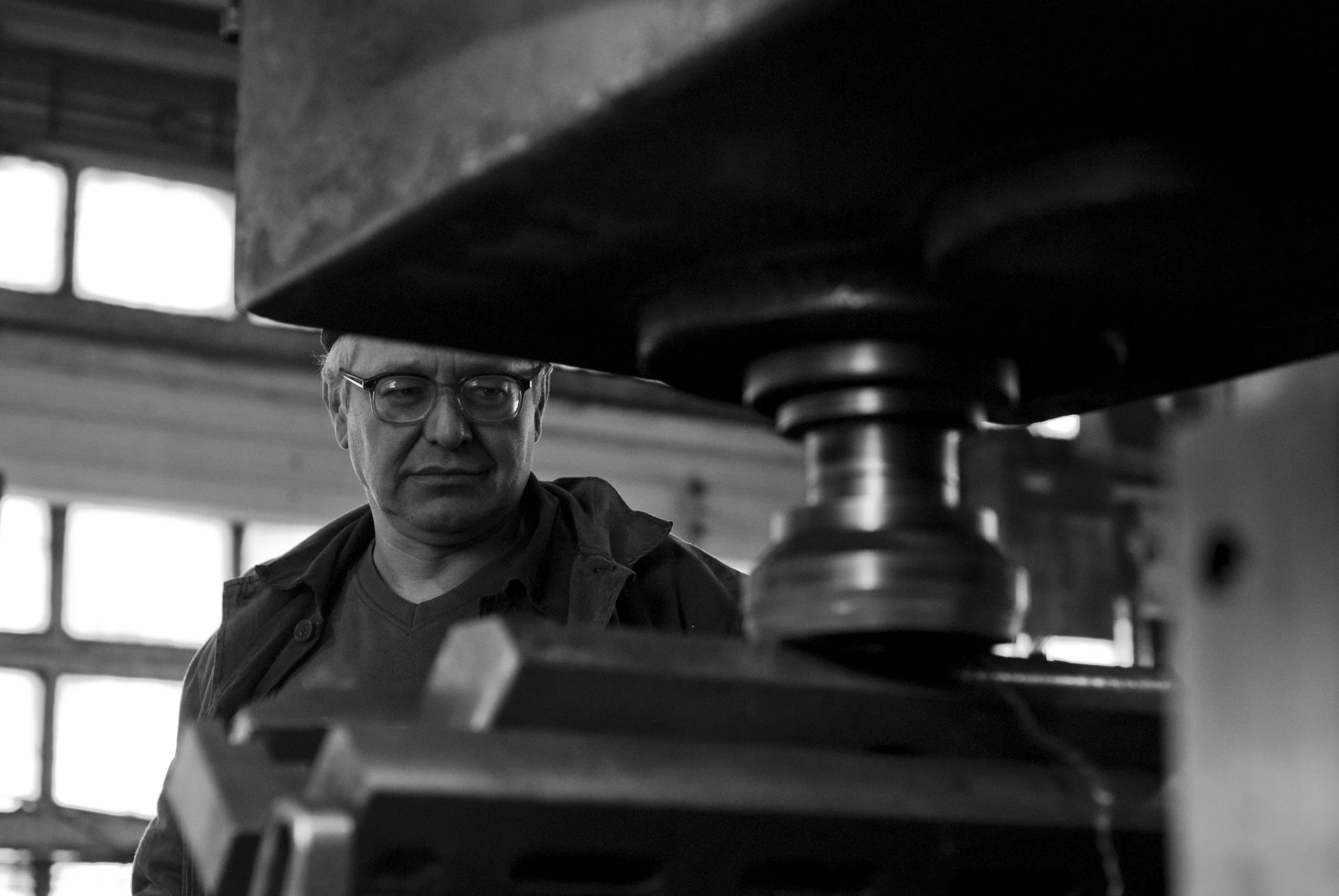 Ein Mann mit Brille und dunkler Kleidung arbeitet unter einer großen Maschine in einer Fabrikhalle.
