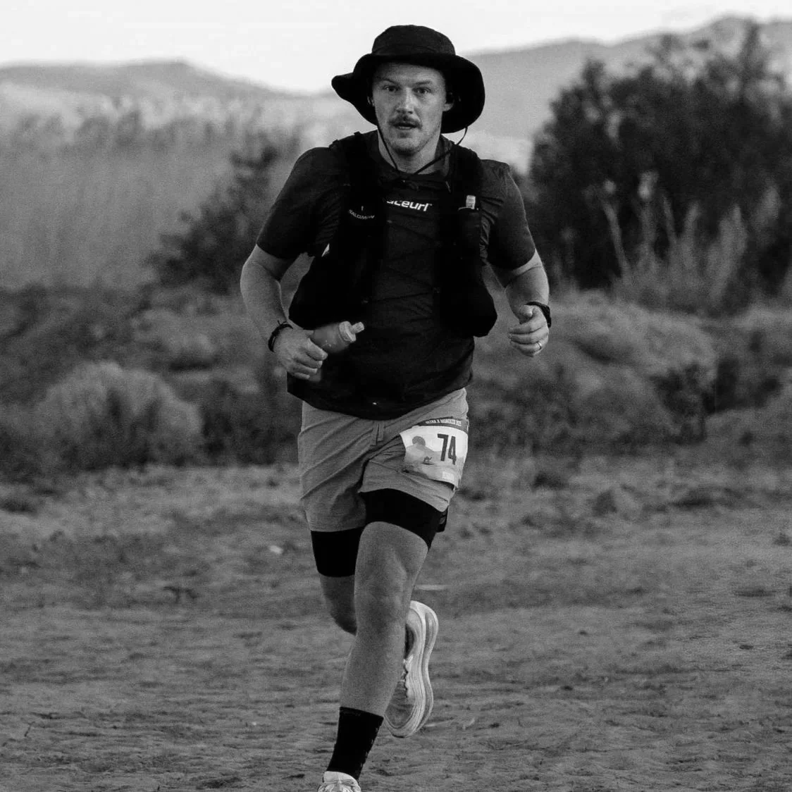 A man running outdoors during a race, wearing a wide-brimmed hat, a dark t-shirt, shorts, and a race number 74 attached to his shorts, with a focused expression.