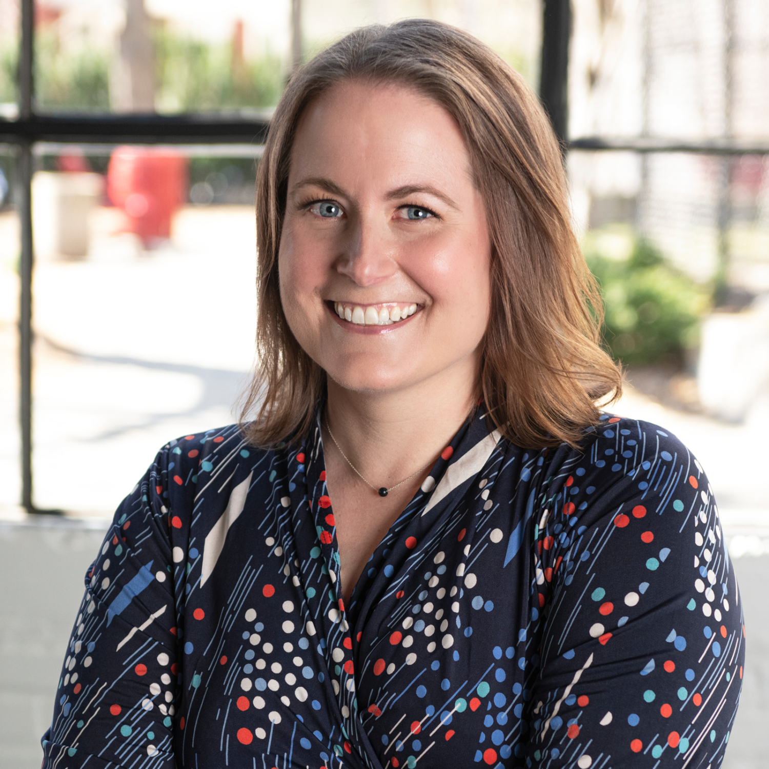 A smiling woman with shoulder-length brown hair, wearing a navy blue blouse with a colorful polka dot and striped pattern, sitting in front of a window with a blurred outdoor background.