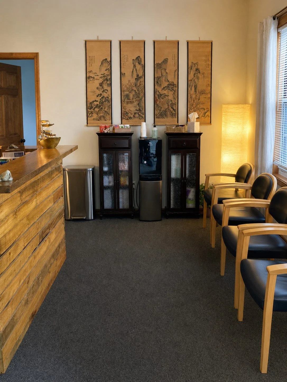 A waiting room with four black chairs with wooden armrests lined up along the right wall, a tall standing lamp with a paper shade near a window, two dark wooden cabinets behind a water cooler, and traditional Asian hanging scroll paintings on the wall.