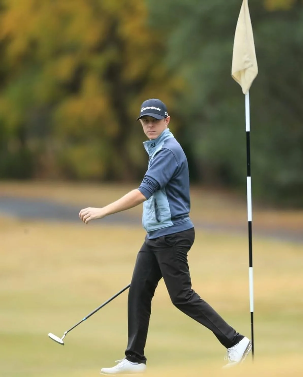 A young male golfer in a blue and gray jacket and black pants on the golf course, with a golf flag behind him.