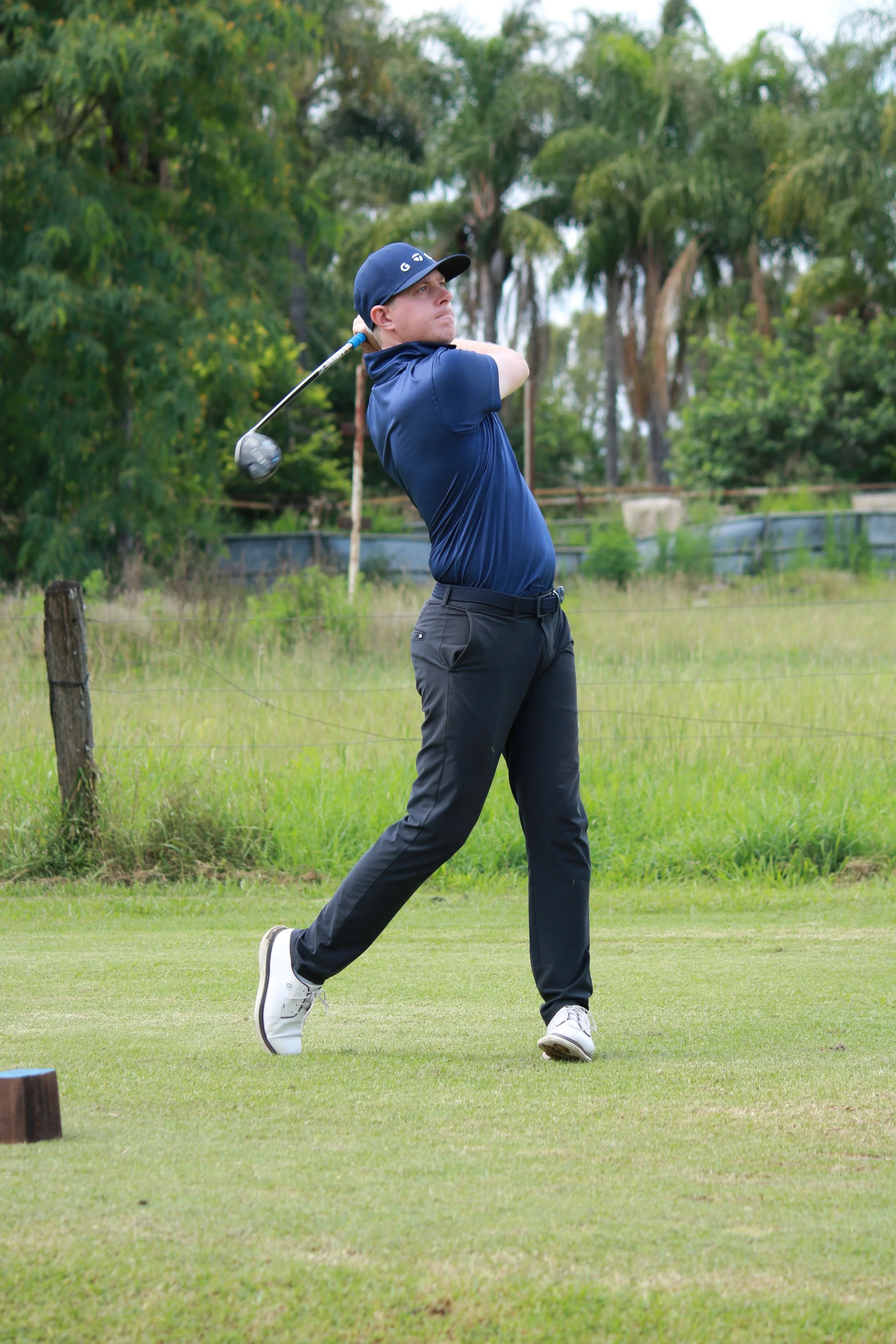 A male golfer swinging a golf club on a green golf course with trees in the background.