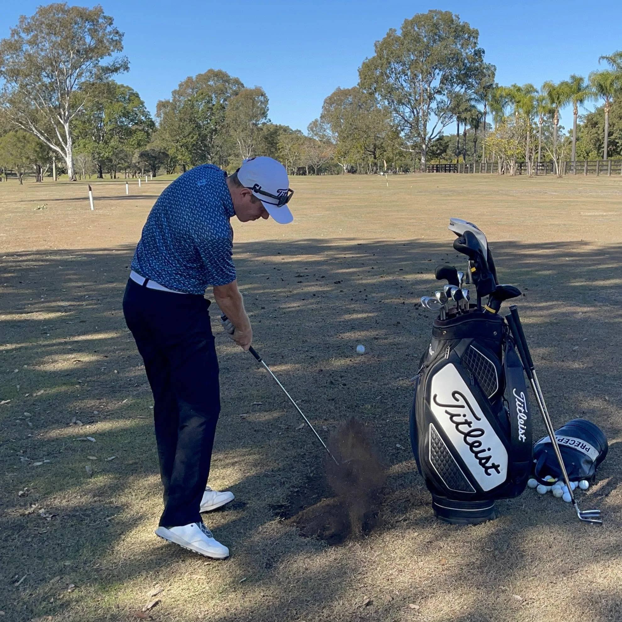 A man preparing to swing a golf club on a golf course with trees and blue sky in the background, with his golf bag and golf balls nearby.