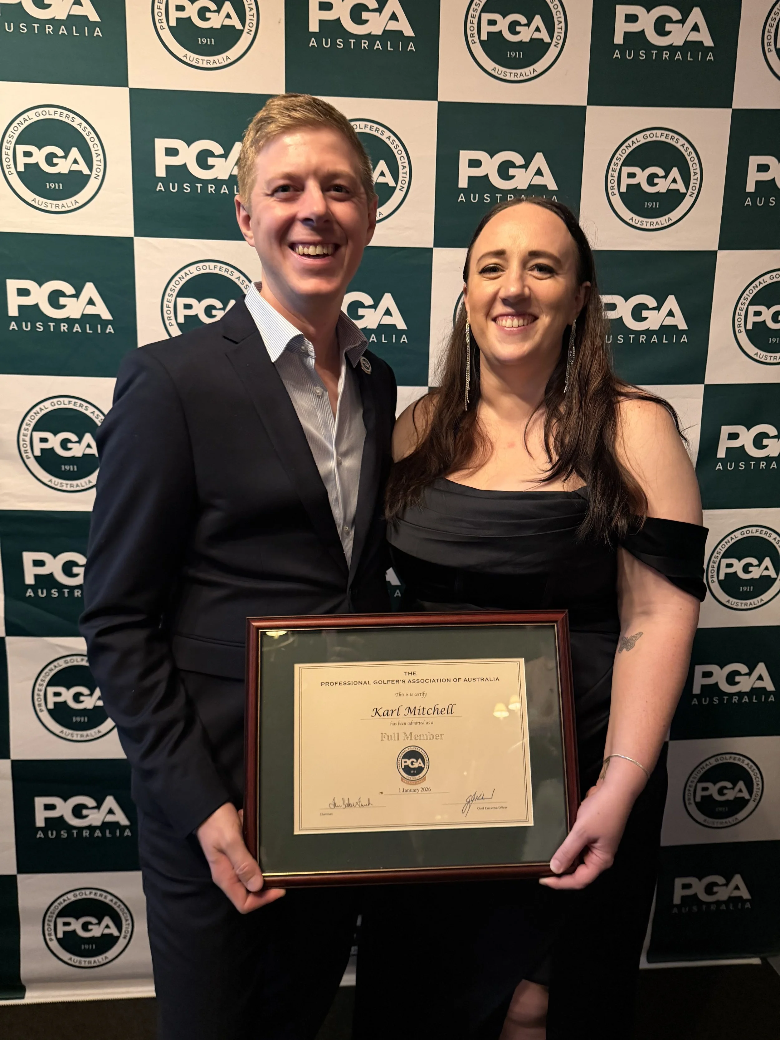 A man and woman smiling and holding a framed certificate at an event with a PGA Australia backdrop.