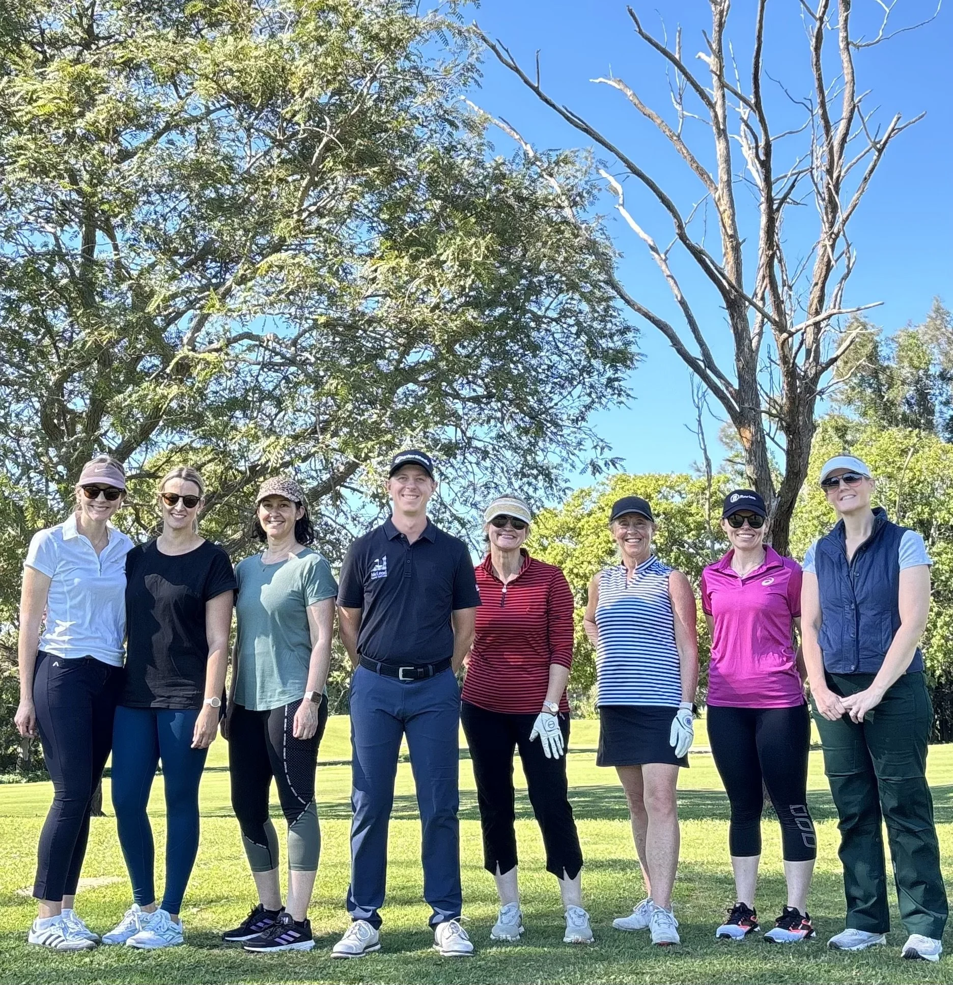 A group of women and one man standing outdoors on a sunny day, dressed in golf attire, some wearing visors and sunglasses, with trees and a grassy area in the background.