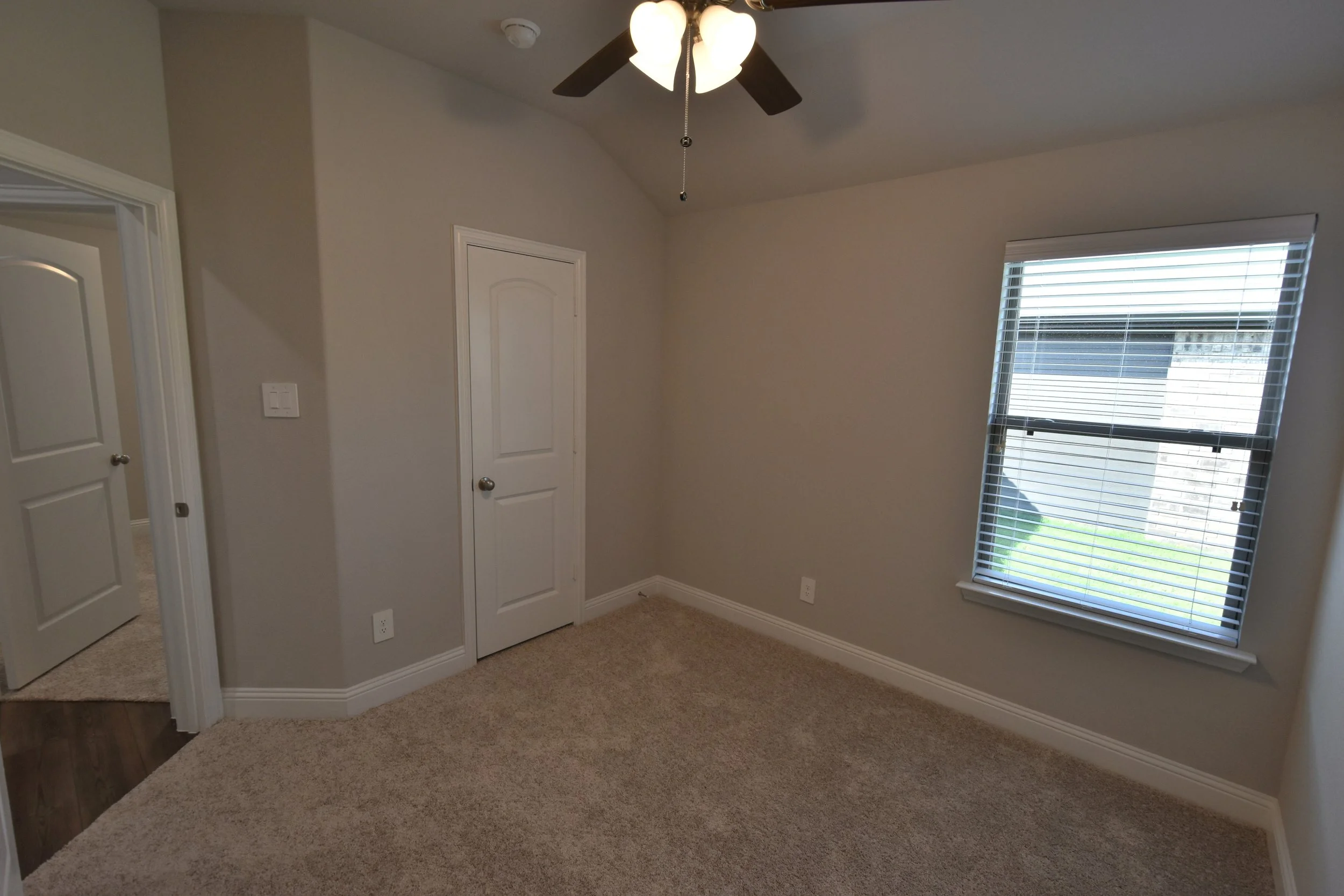 A corner of a room with beige carpet, beige walls, and a white ceiling. There is a window with closed blinds on the right side, and two white doors with silver knobs on the left side. A ceiling fan with a light fixture is mounted on the ceiling.