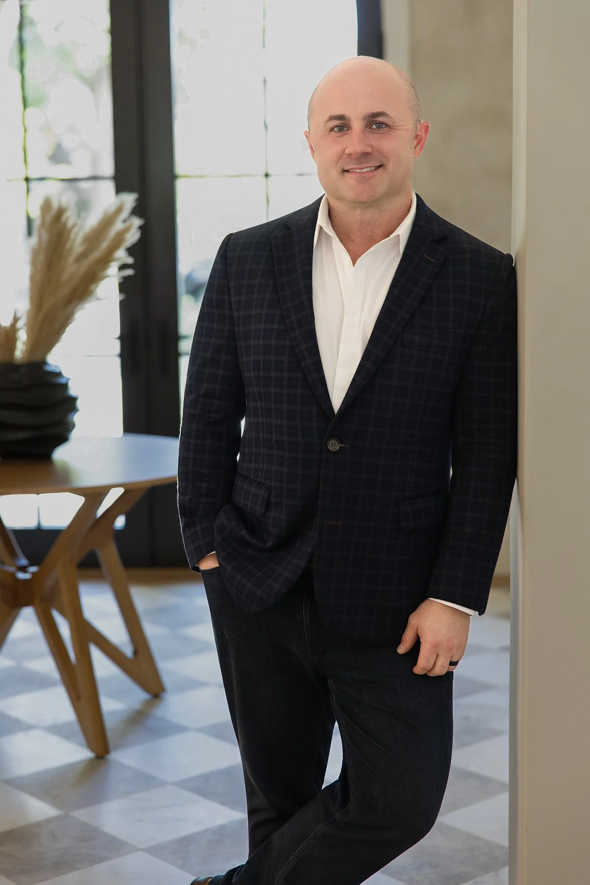 A man in a dark checkered blazer and white shirt stands smiling indoors near a wall, with a dining table and window in the background.