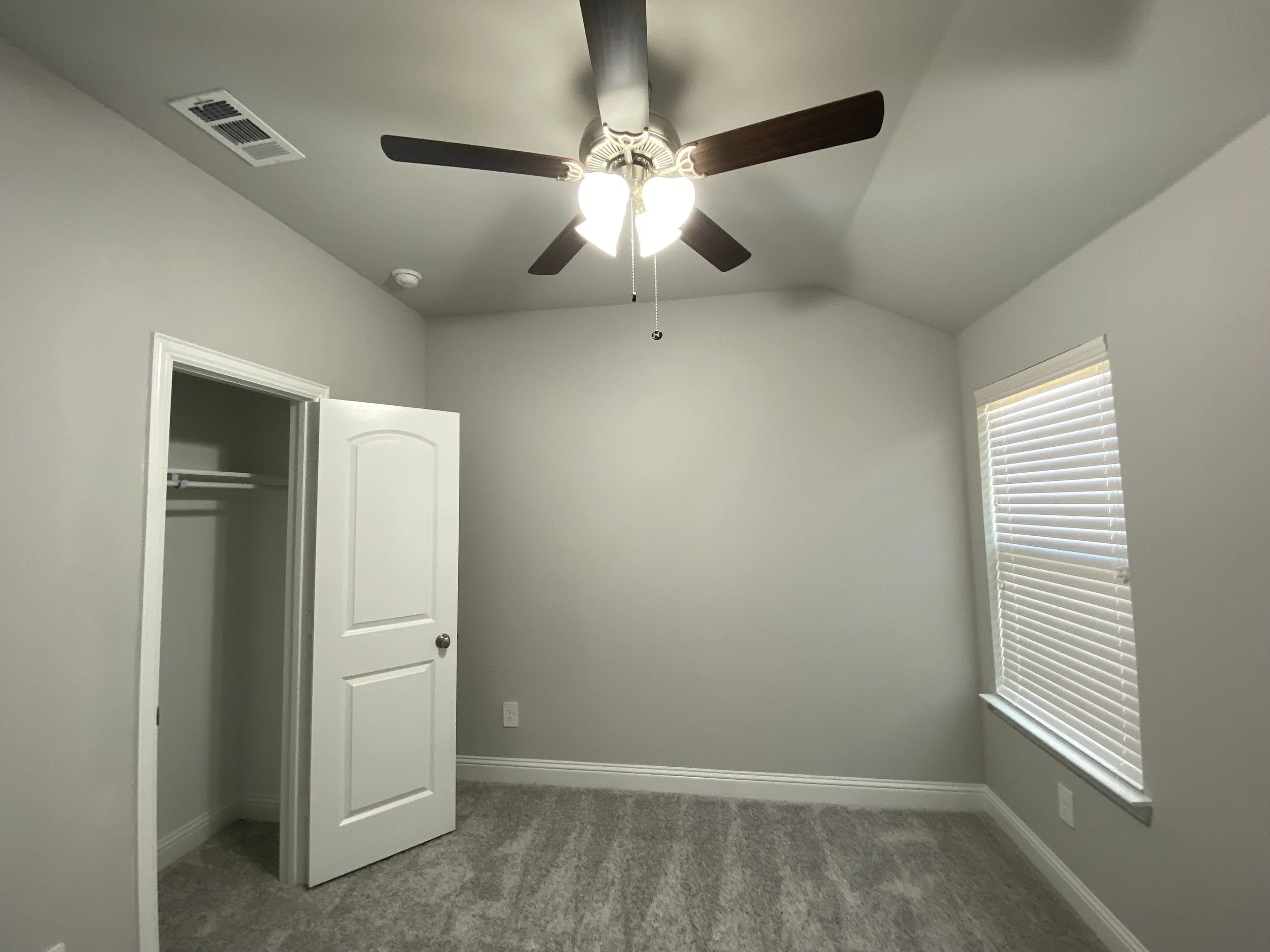 Empty room with gray walls, a white ceiling fan with light, a window with blinds, a closet door with a white frame, and carpeted flooring.