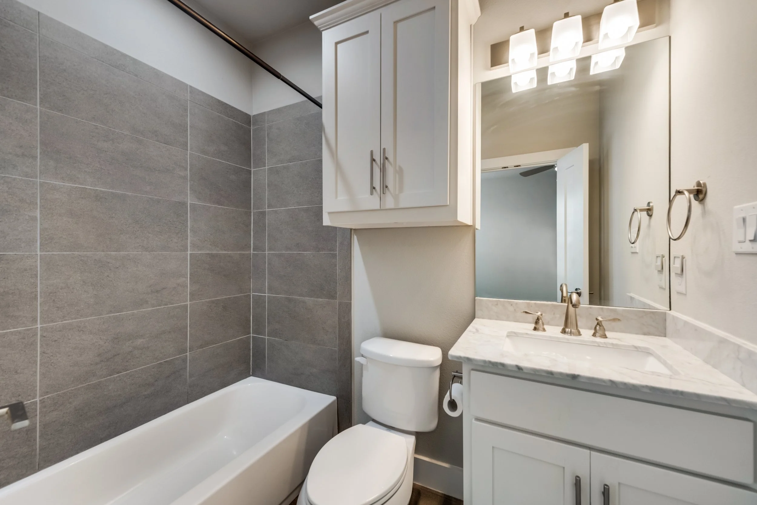 A modern bathroom with a bathtub, gray tiled wall, white cabinet, mirror, and a marble countertop with a sink and faucet.