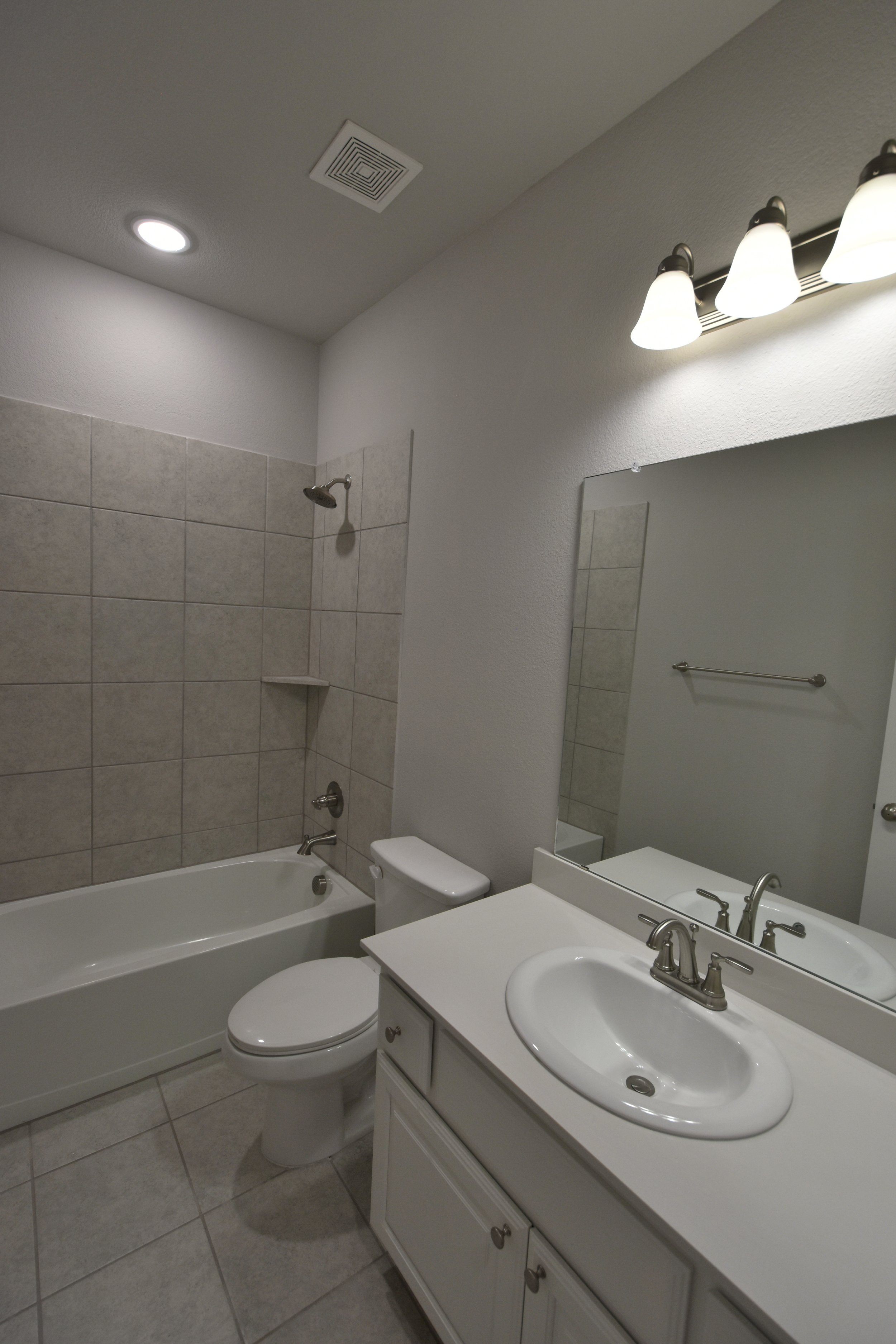 Bathroom with bathtub, toilet, mirror, sink, and vanity with cabinet, tiled wall, and ceiling lights.