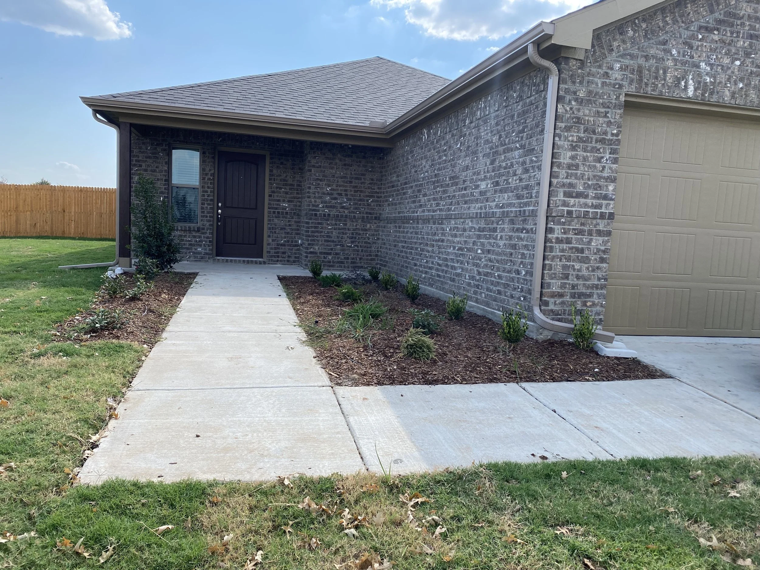 Front view of a house with a brick exterior, a black front door, a concrete walkway, and a closed garage door. There is a small garden with bushes and a lawn, fenced by a wooden fence, under a partly cloudy sky.