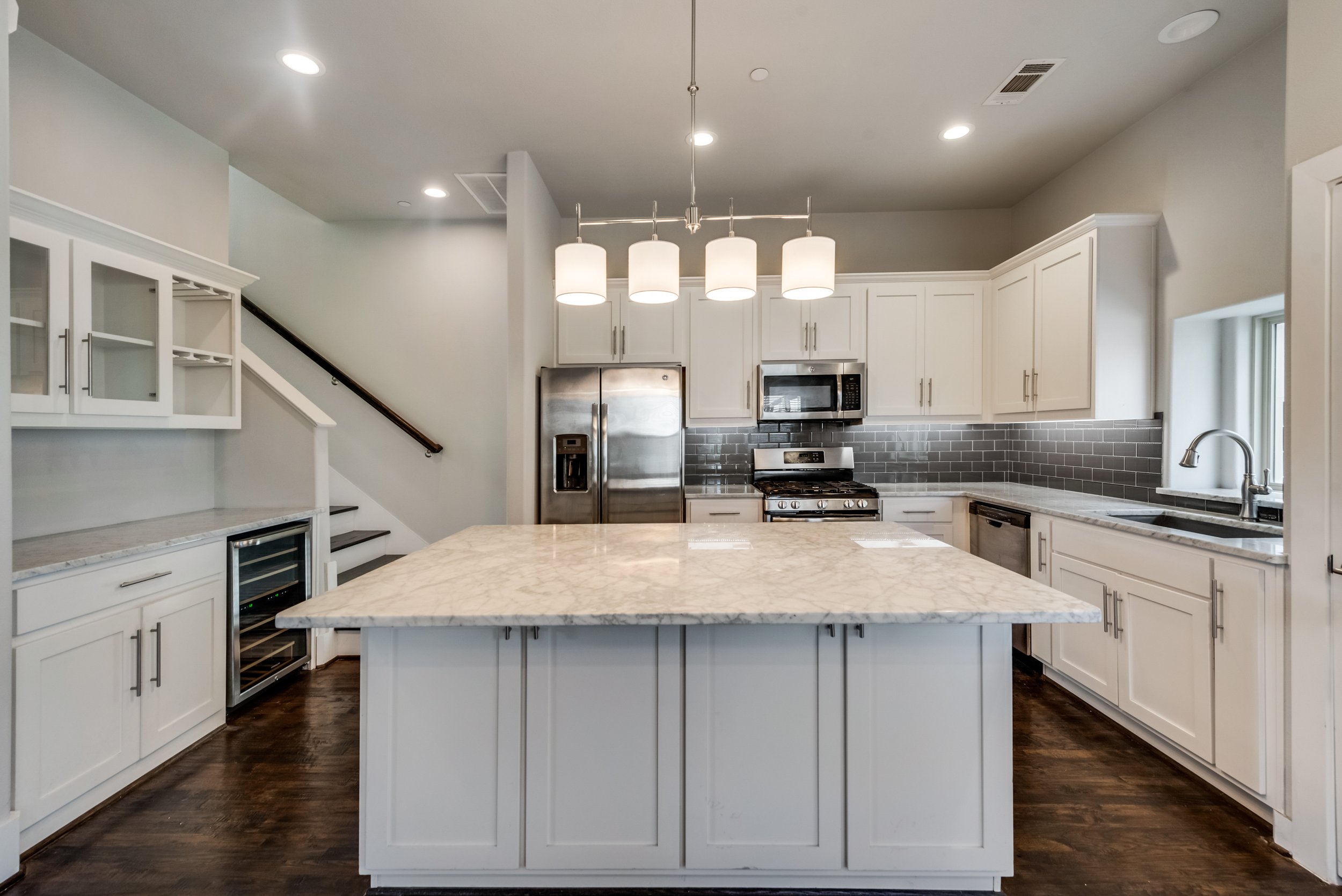 Modern kitchen with white cabinets, stainless steel appliances, marble island, gray subway tile backsplash, dark hardwood floors, and a contemporary light fixture with five white pendant lamps.