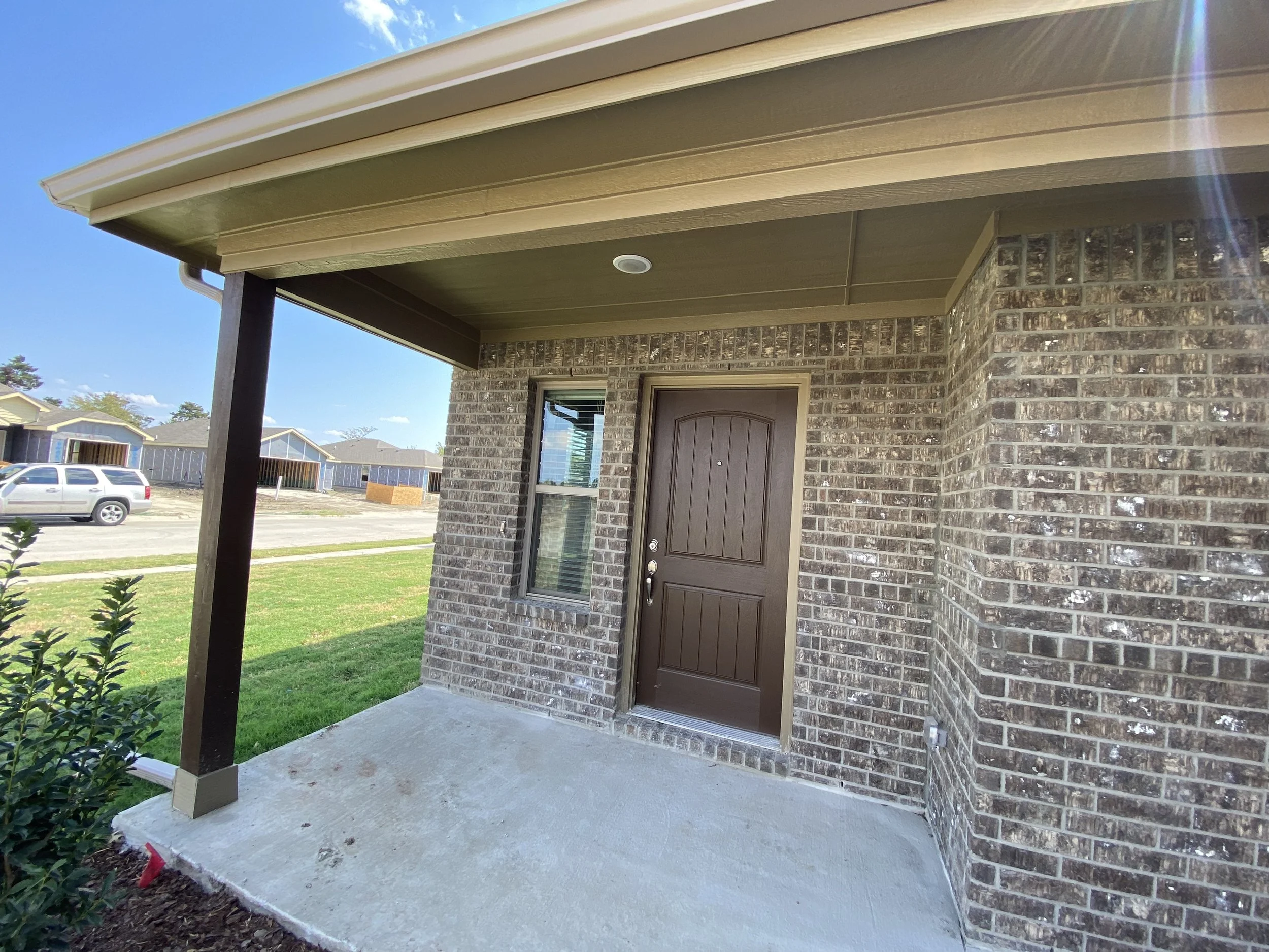 Front porch of a brick house with a brown door and a window, under a covered roof, with a concrete base and green grass around it.