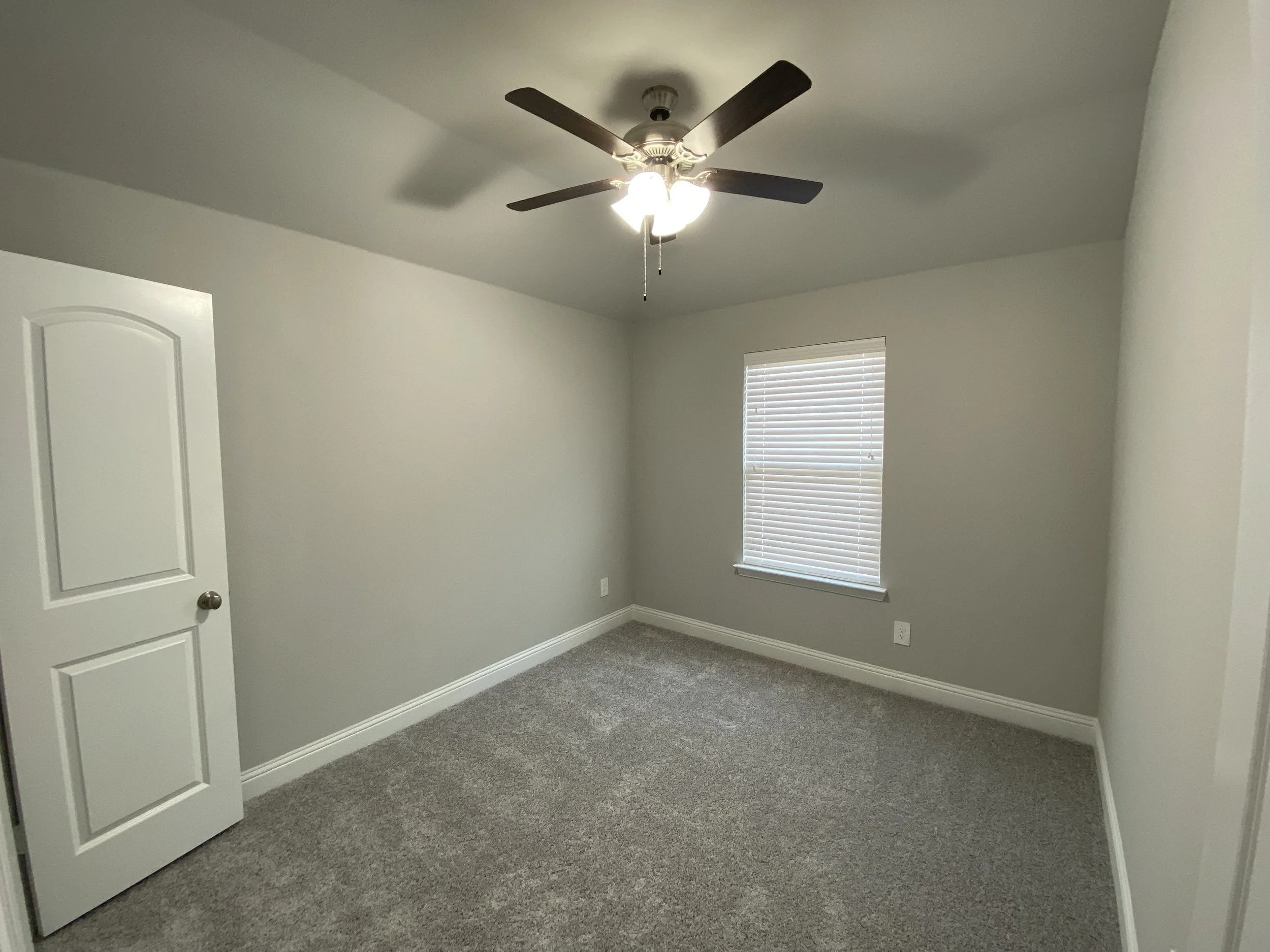 Empty room with beige walls, a ceiling fan with lights, a window with blinds, and gray carpet flooring.