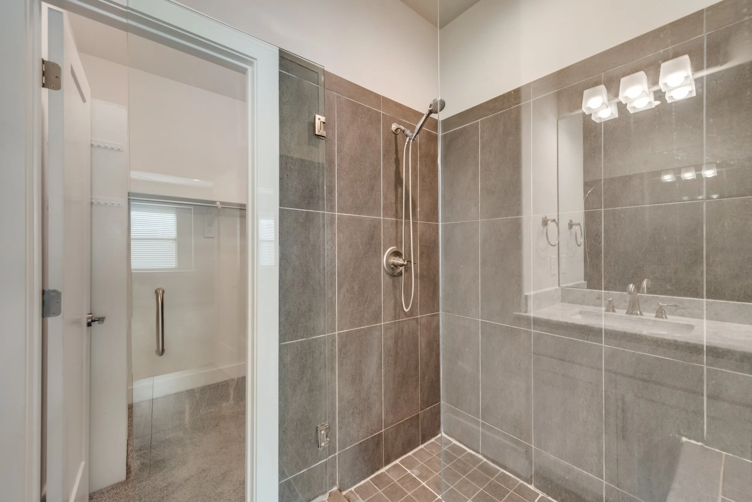 Bathroom with tiled walls and floor, a mirror, a light fixture, a shower with a handheld showerhead, and a door with a handle.