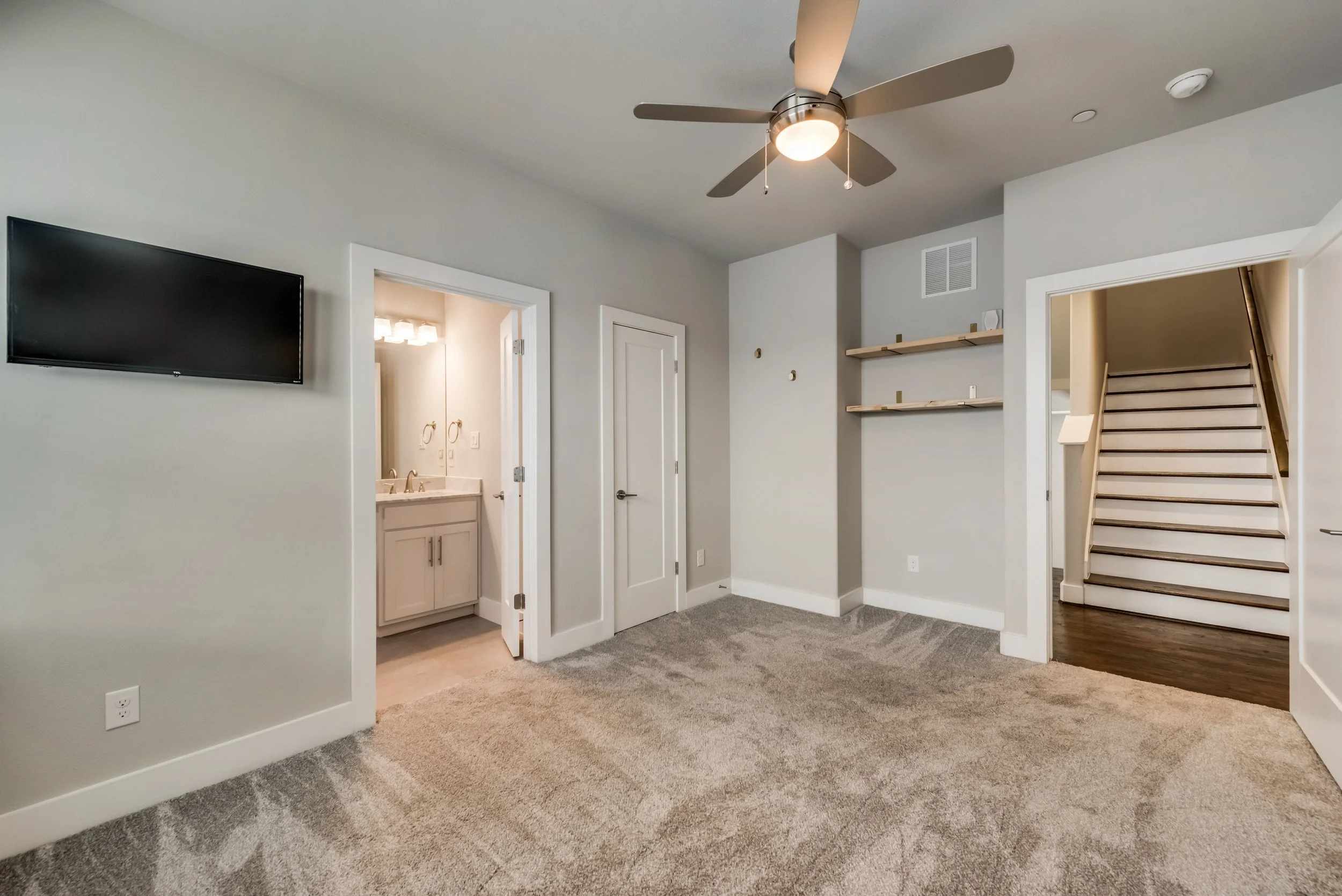 Interior view of a room with beige carpet, white walls, a mounted flat-screen TV, and an open doorway leading to a bathroom with a white vanity and sink. To the right, there are two wooden shelves on the wall and a staircase with wooden steps and whi