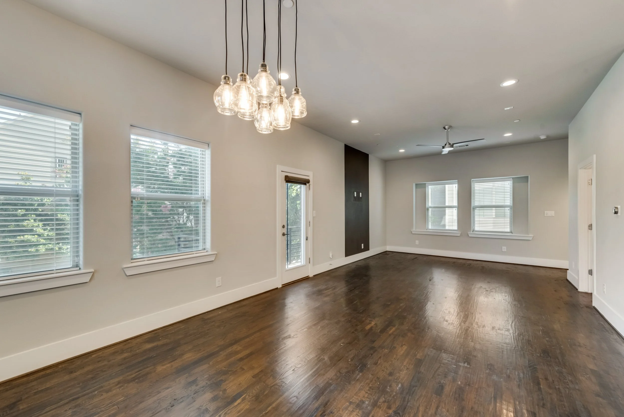 Empty living room with three large windows with white blinds, a door with glass panel, dark wood flooring, white walls, and a modern chandelier with multiple glass bulb covers hanging from the ceiling.