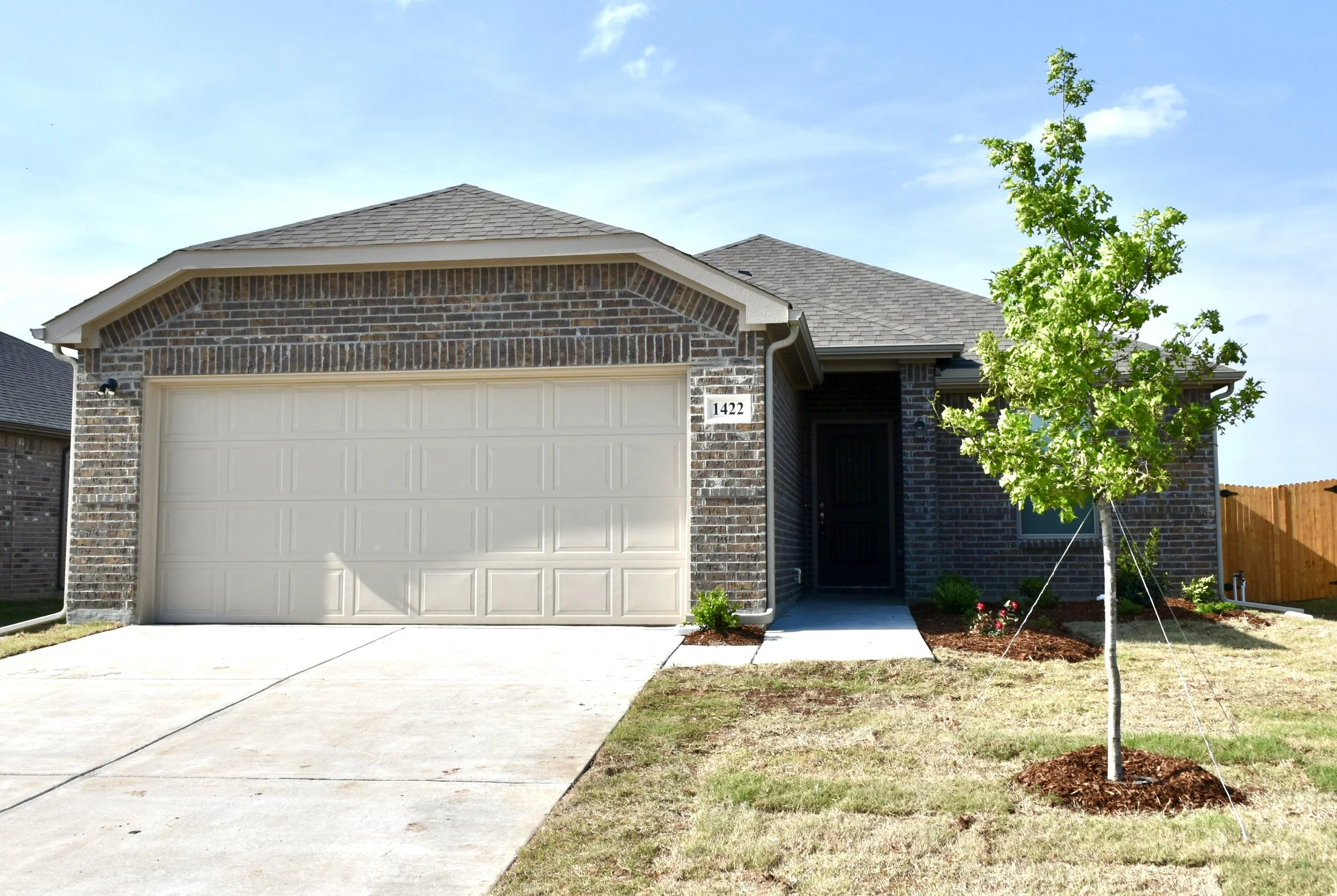 Front view of a brick house with a white garage door, a small front porch, a young tree, and a well-maintained lawn, under a partly cloudy blue sky.