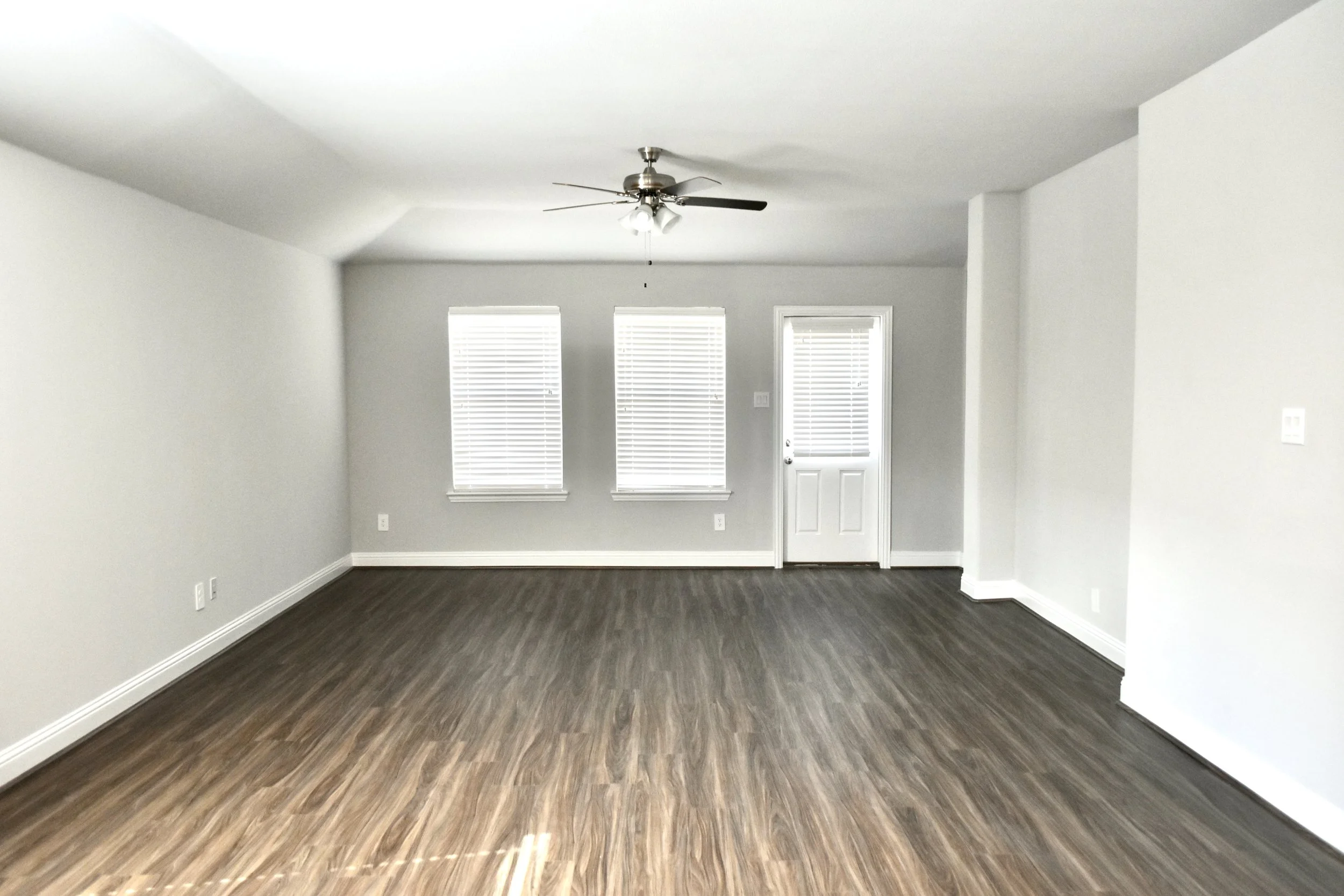Empty living room with white walls, dark wood flooring, three windows with blinds, and a ceiling fan with light fixtures.