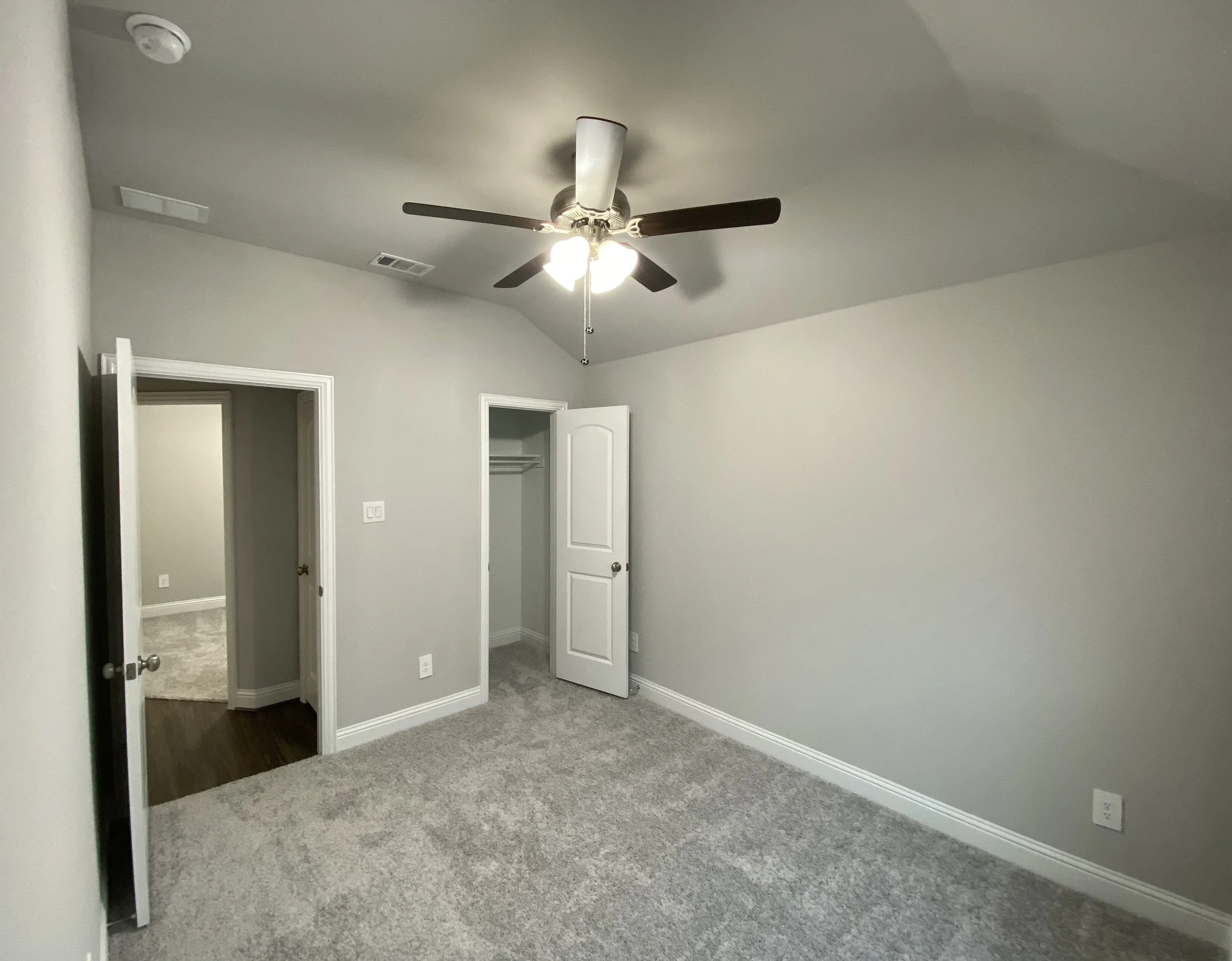 Empty bedroom with gray walls, a ceiling fan with lights, a door slightly open, and two open closet doors.