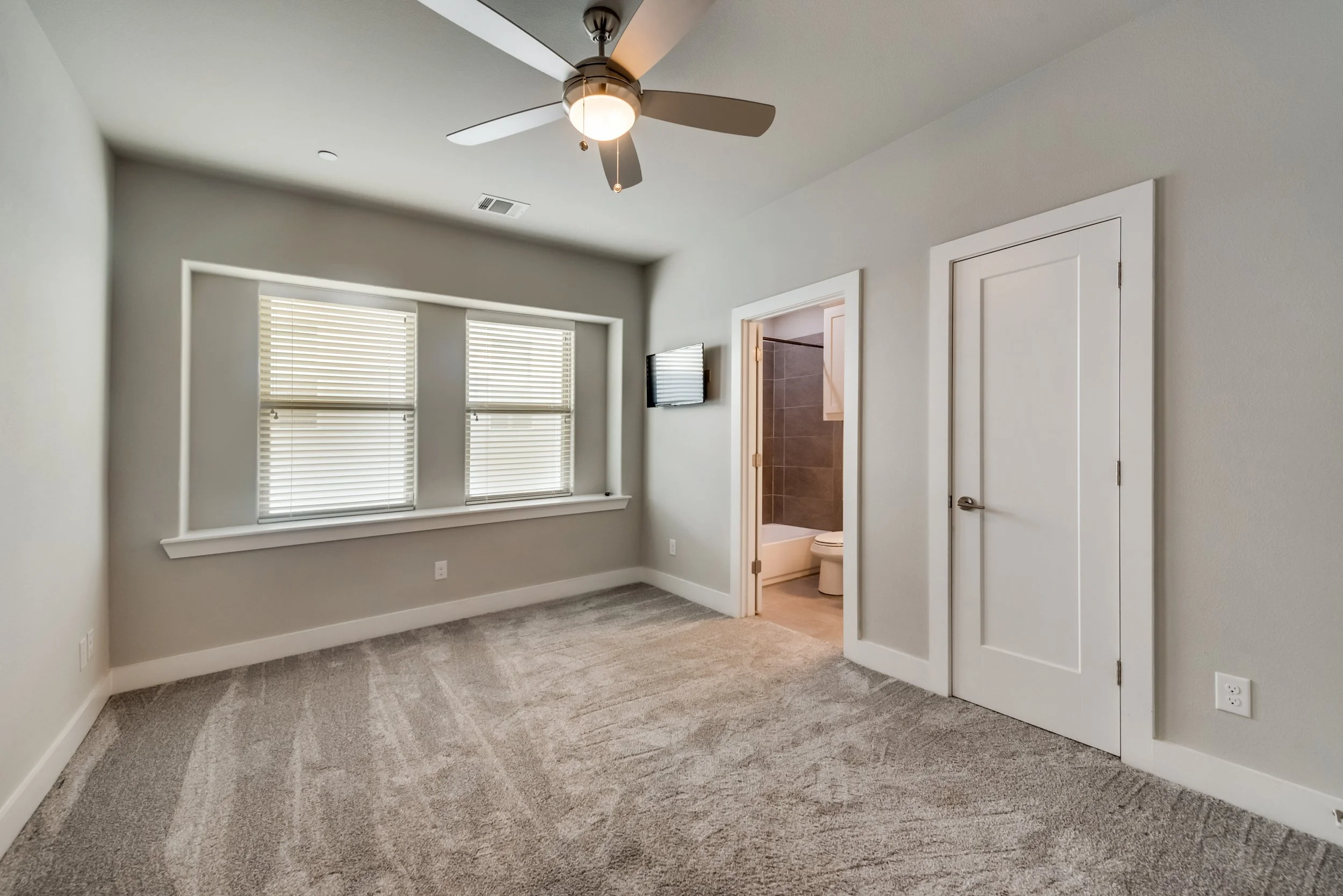 Empty bedroom with beige carpet, white walls, two windows with blinds, a ceiling fan with light, and a door leading to a bathroom with a toilet.