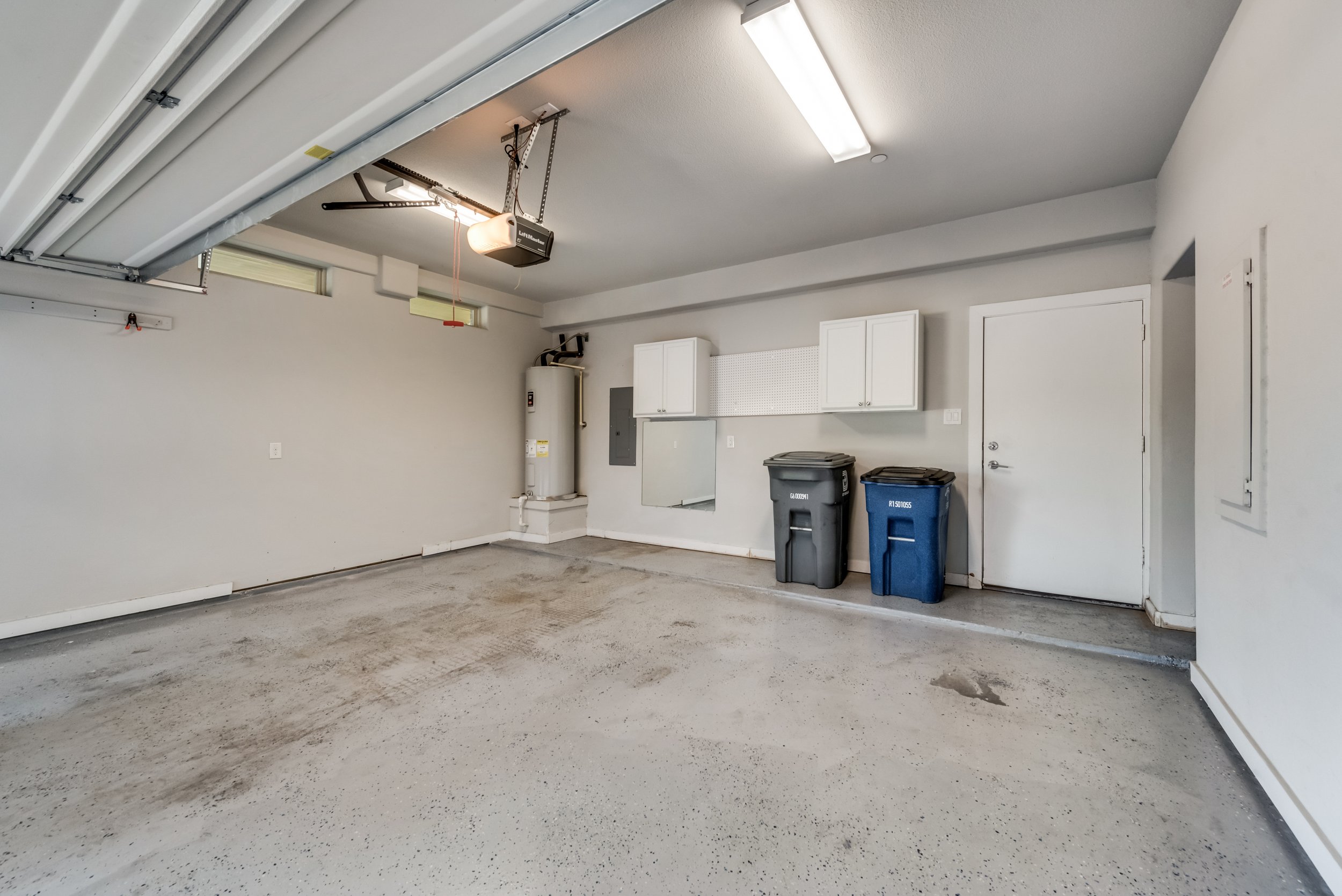 Empty garage with concrete floor, white walls, a garage door partially open, and two trash bins near a door.
