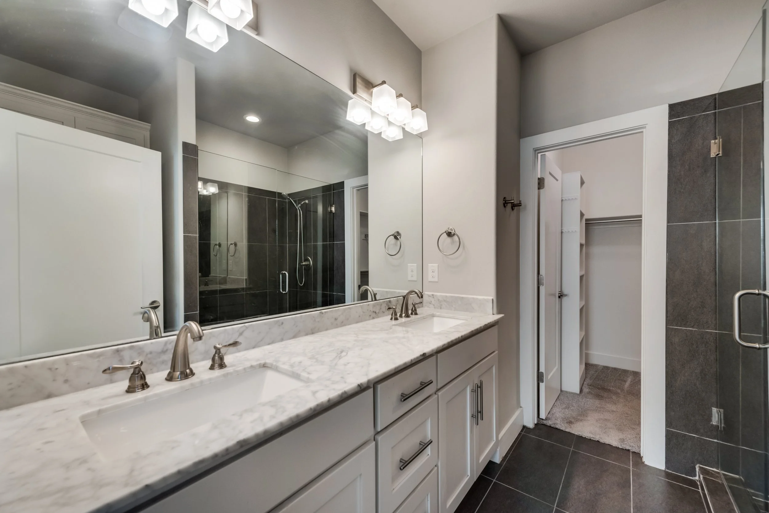 Modern bathroom with large mirror, double sink vanity with marble countertop, dark tiled walk-in shower, towel ring, and an open doorway to storage closet.