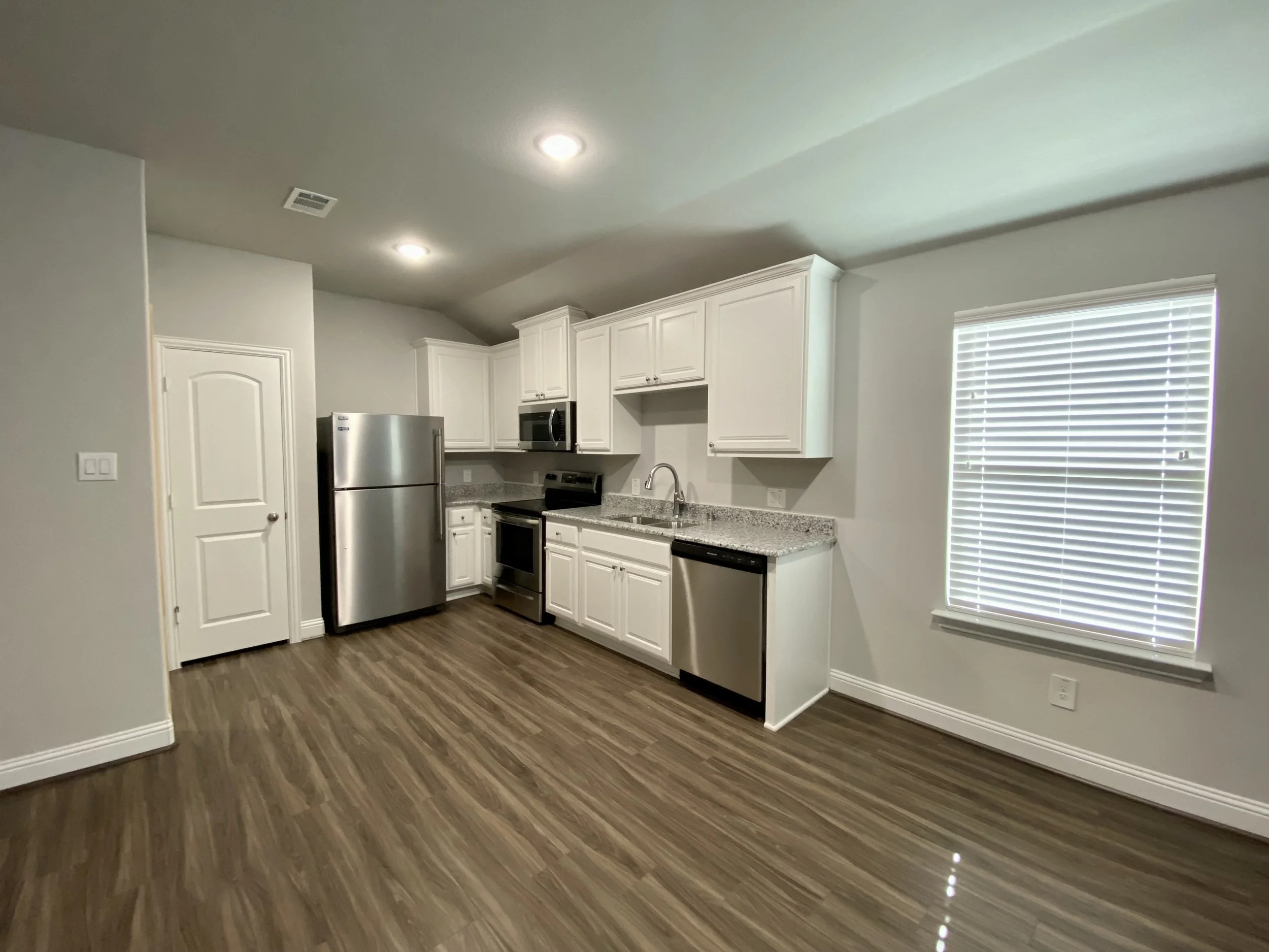 Kitchen with white cabinets, stainless steel appliances including a refrigerator, microwave, and stove, with granite countertops and wood-look flooring, a window with blinds, and recessed ceiling lights.