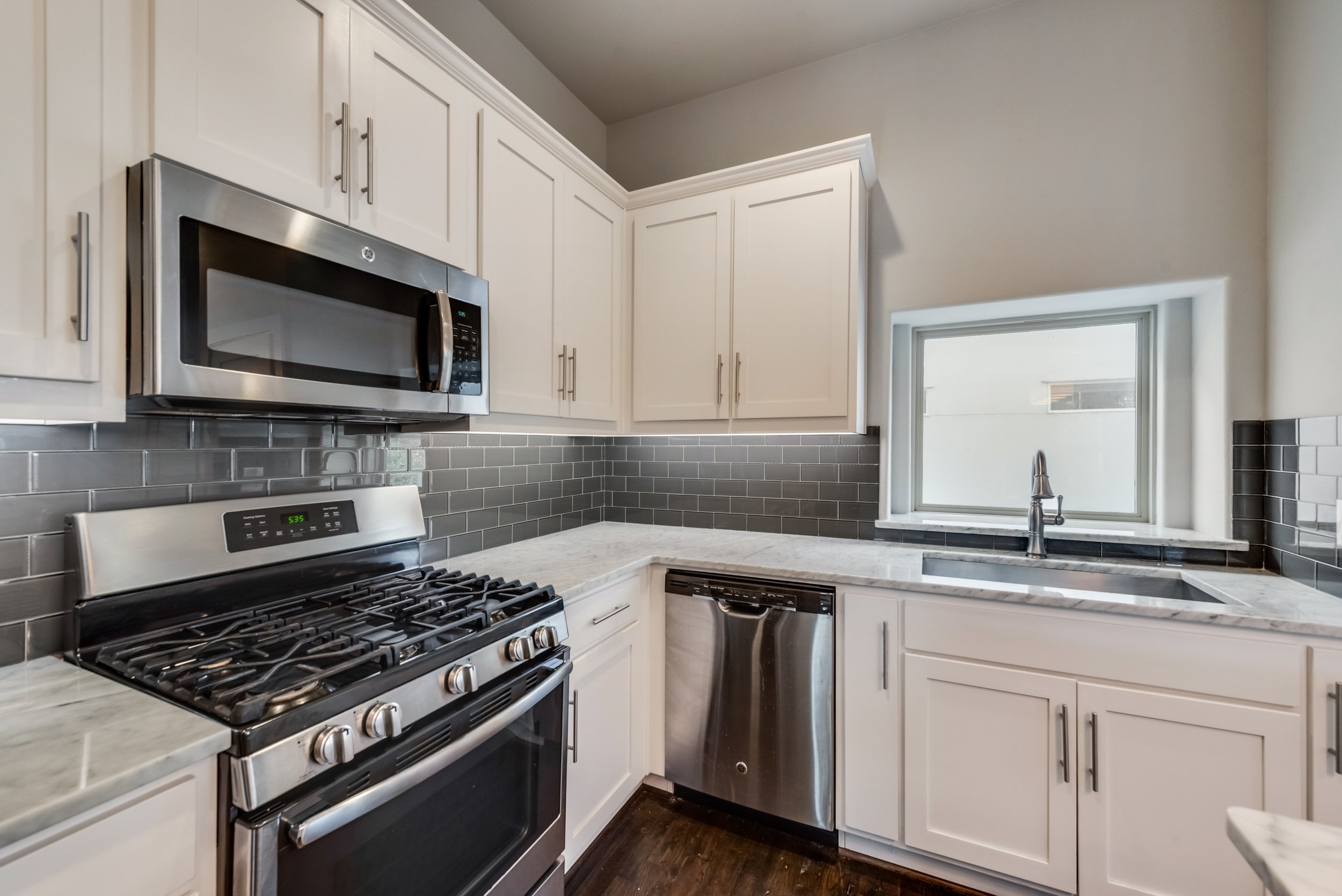 Kitchen with white cabinets, stainless steel microwave, gas stove, gray subway tile backsplash, and a small window over the sink.