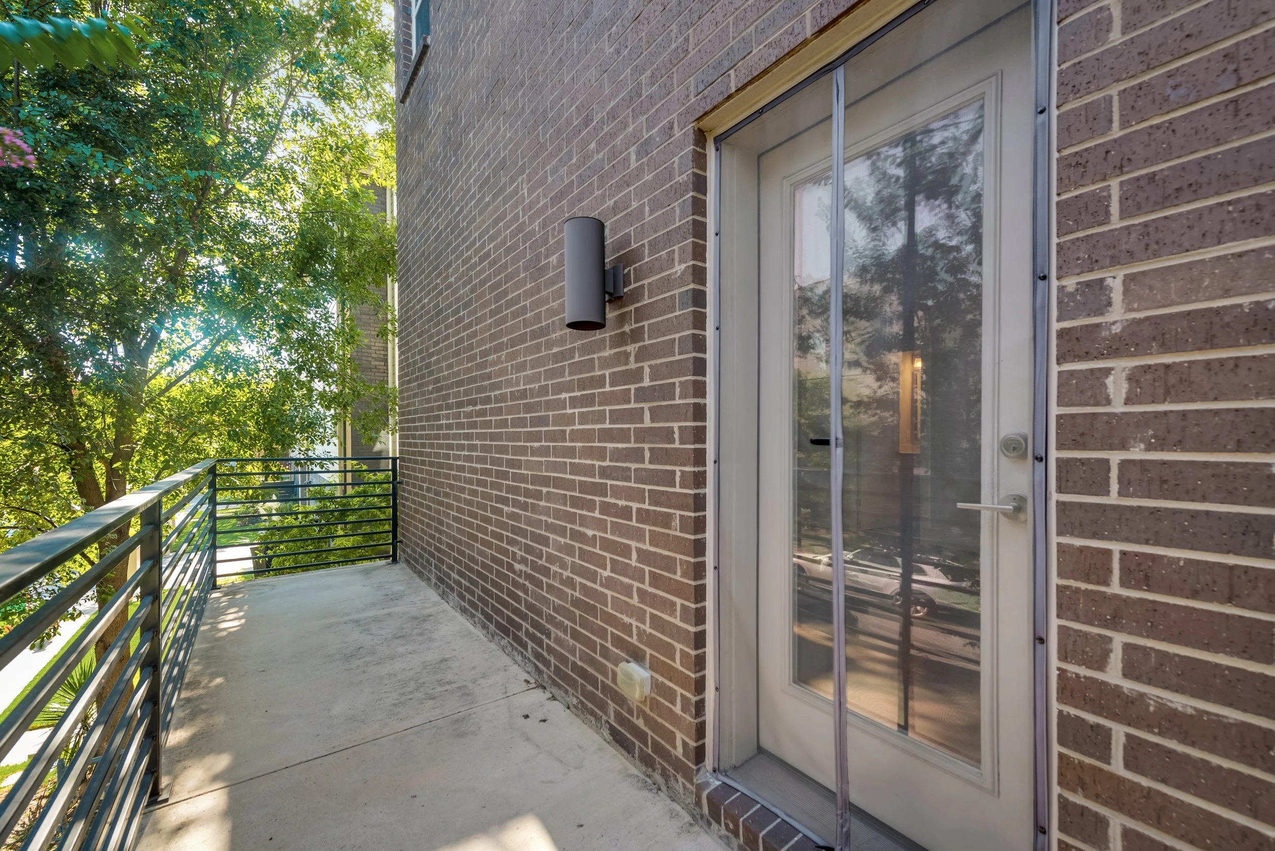 A balcony with a metal railing, concrete floor, brick wall, and a door with a glass window, surrounded by greenery and trees.