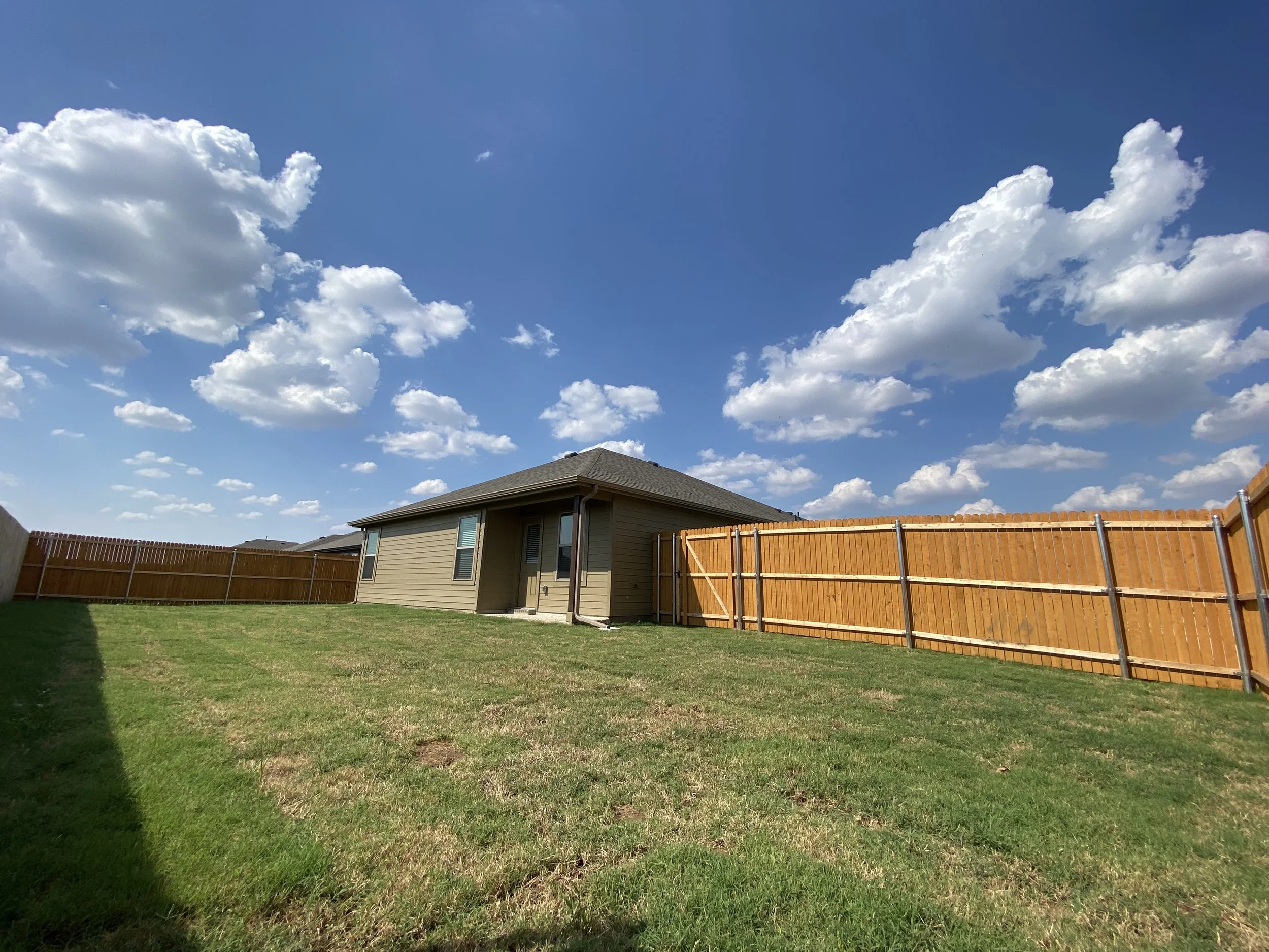 A backyard with green grass, a wooden fence, a house with beige siding, and a blue sky with white clouds.